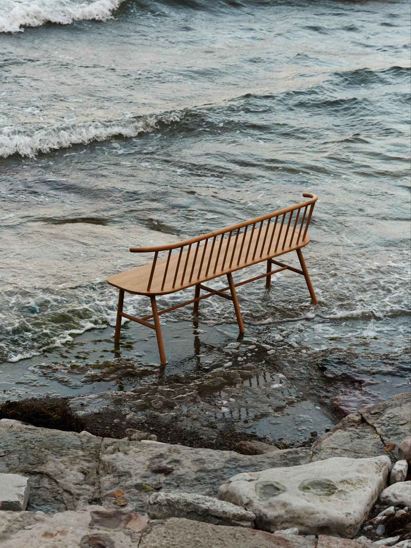 Jasna, drewniana ławka ze szczebelkami i wygiętym oparciem stoi w płytkiej wodzie na skalistej plaży z delikatnymi falami.