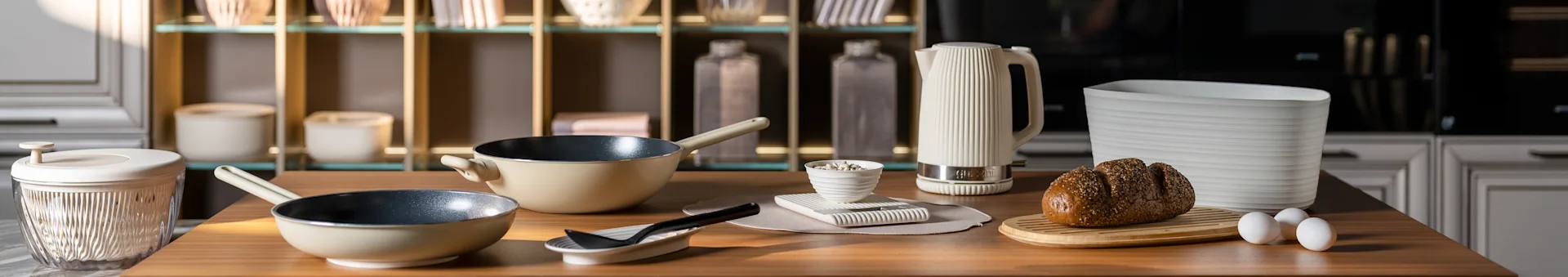 Blat kuchenny z beżowymi patelniami, białym żebrowanym czajnikiem, chlebem z ziarnami na desce i jajkami. Nowoczesne akcesoria kuchenne.