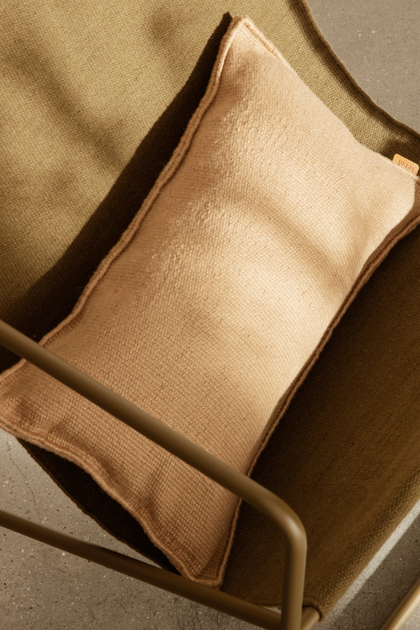 Widok z góry na jasnobrązową poduszkę na ciemnozielonym teksturowanym fotelu z tkaniny, z częścią metalowej ramy i betonową podłogą.