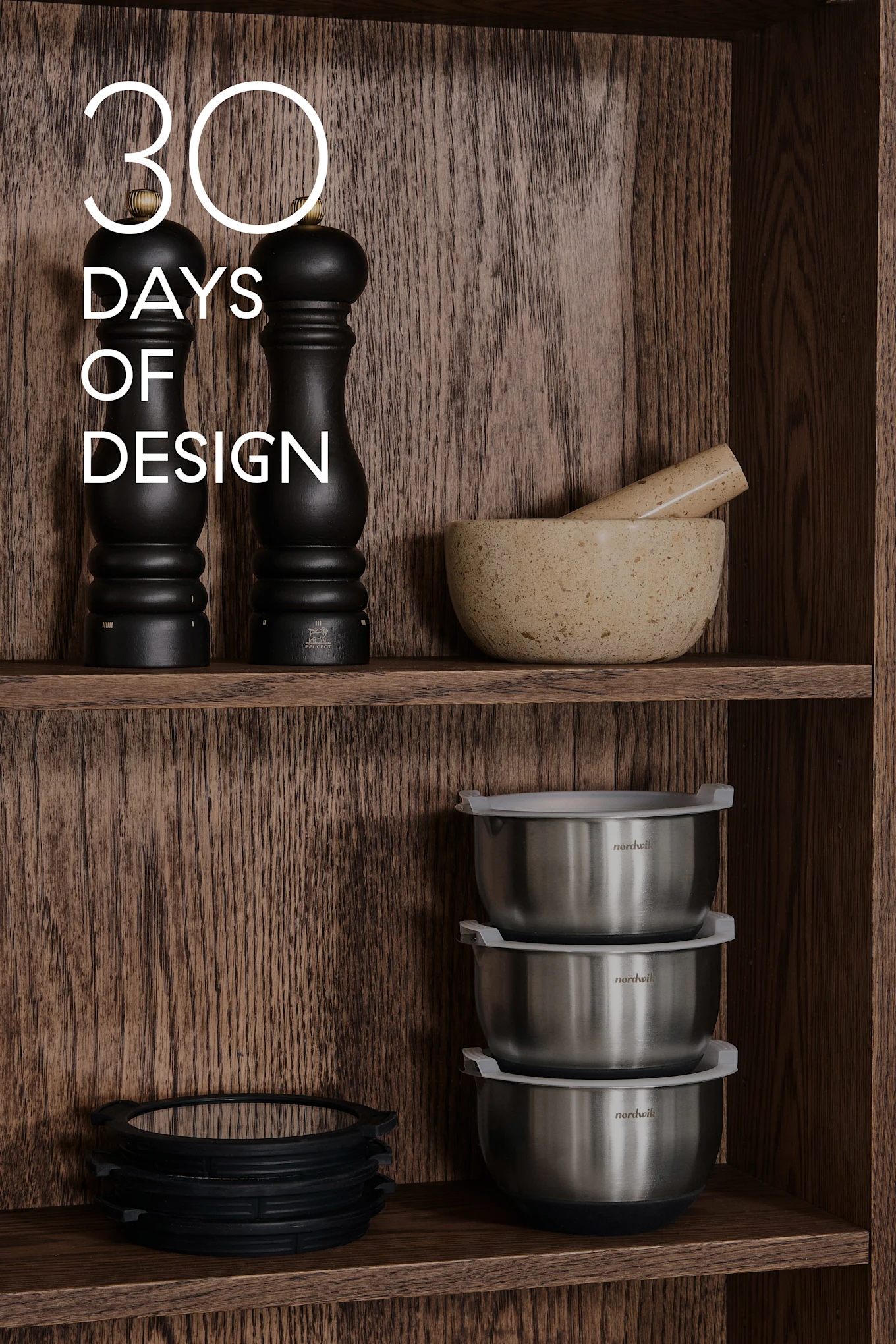 Drewniane półki z przyborami kuchennymi: czarne młynki, moździerz i tłuczek, stalowe miski, silikonowe pierścienie. Tekst: 30 DAYS OF DESIGN.