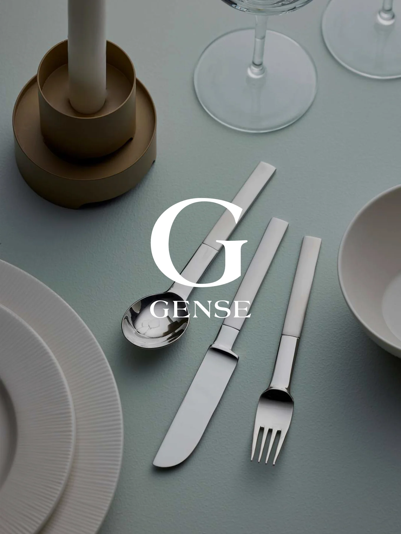 Nakrycie stołu ze srebrnym zestawem sztućców Gense (łyżka, nóż, widelec), białymi talerzami, świecą i szklankami na jasnoniebieskiej powierzchni.