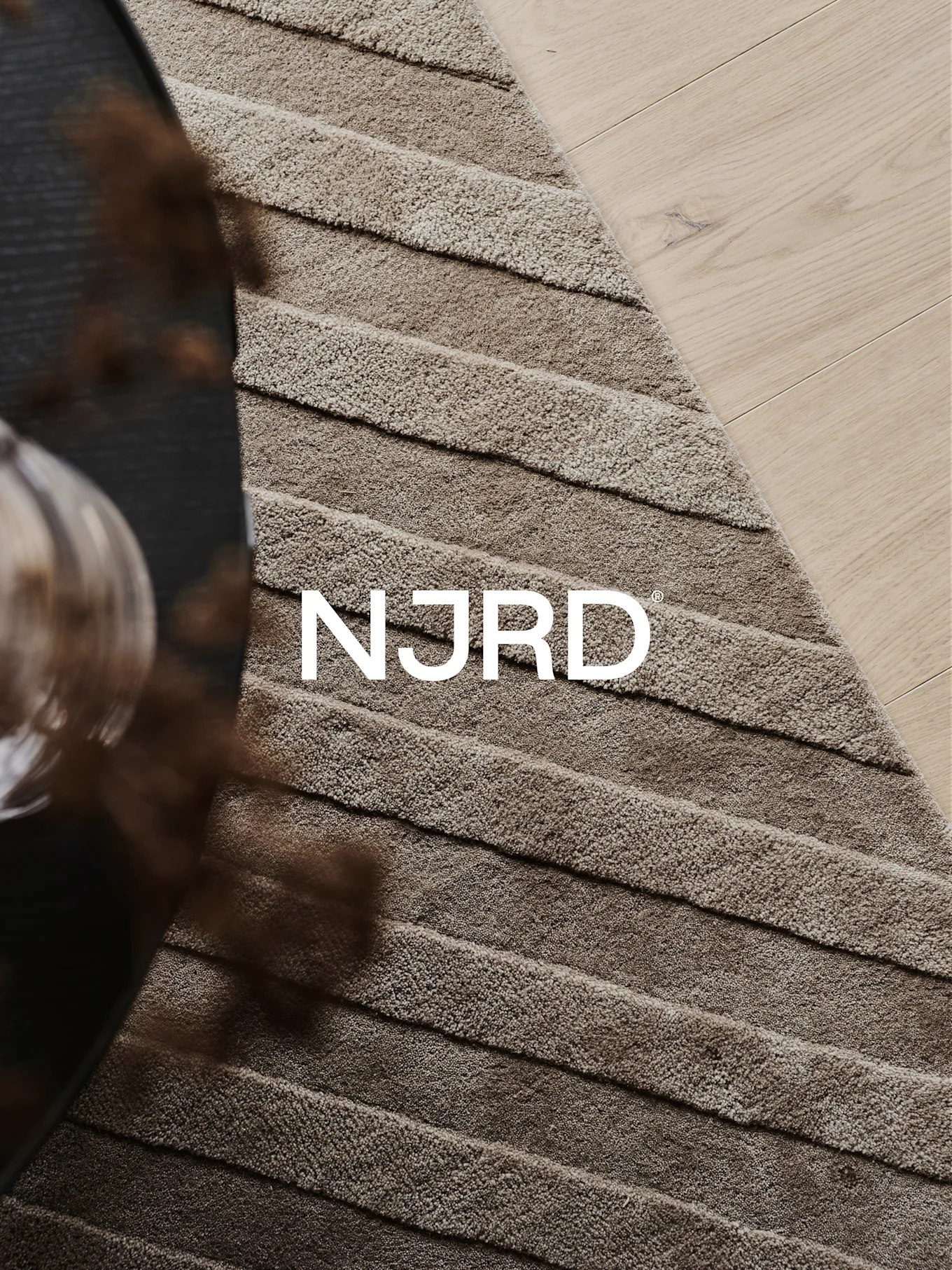 Zbliżenie na jasnobrązowy dywan shaggy z poziomymi grzbietami, częściowo na jasnej drewnianej podłodze, z białym logo 'NJRD'.