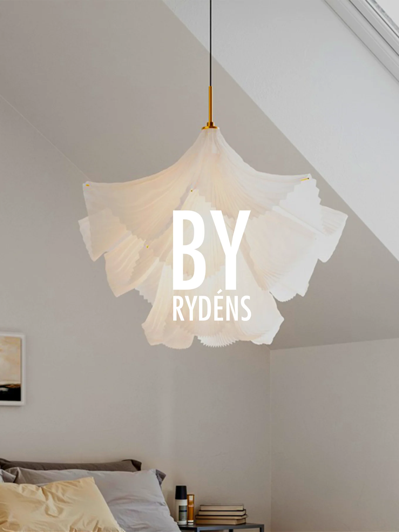 Biała, plisowana rzeźbiarska lampa wisząca wisi nad łóżkiem z poduszkami i stolikiem nocnym. Napis 'BY RYDÉNS' jest nałożony na lampę.