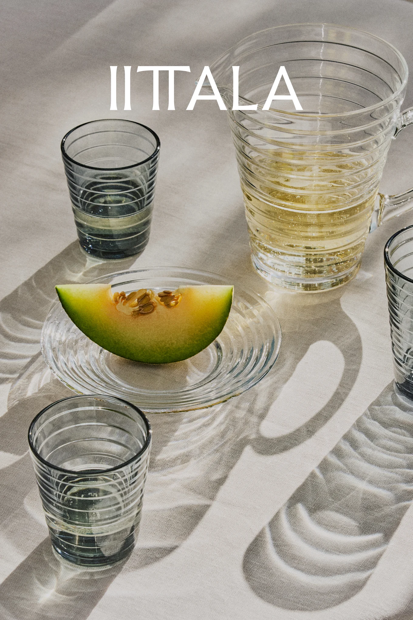 Logo IITTALA nad szklankami, dzbanek z napojem gazowanym i plasterek melona na talerzu, wszystko na jasnym materiale.