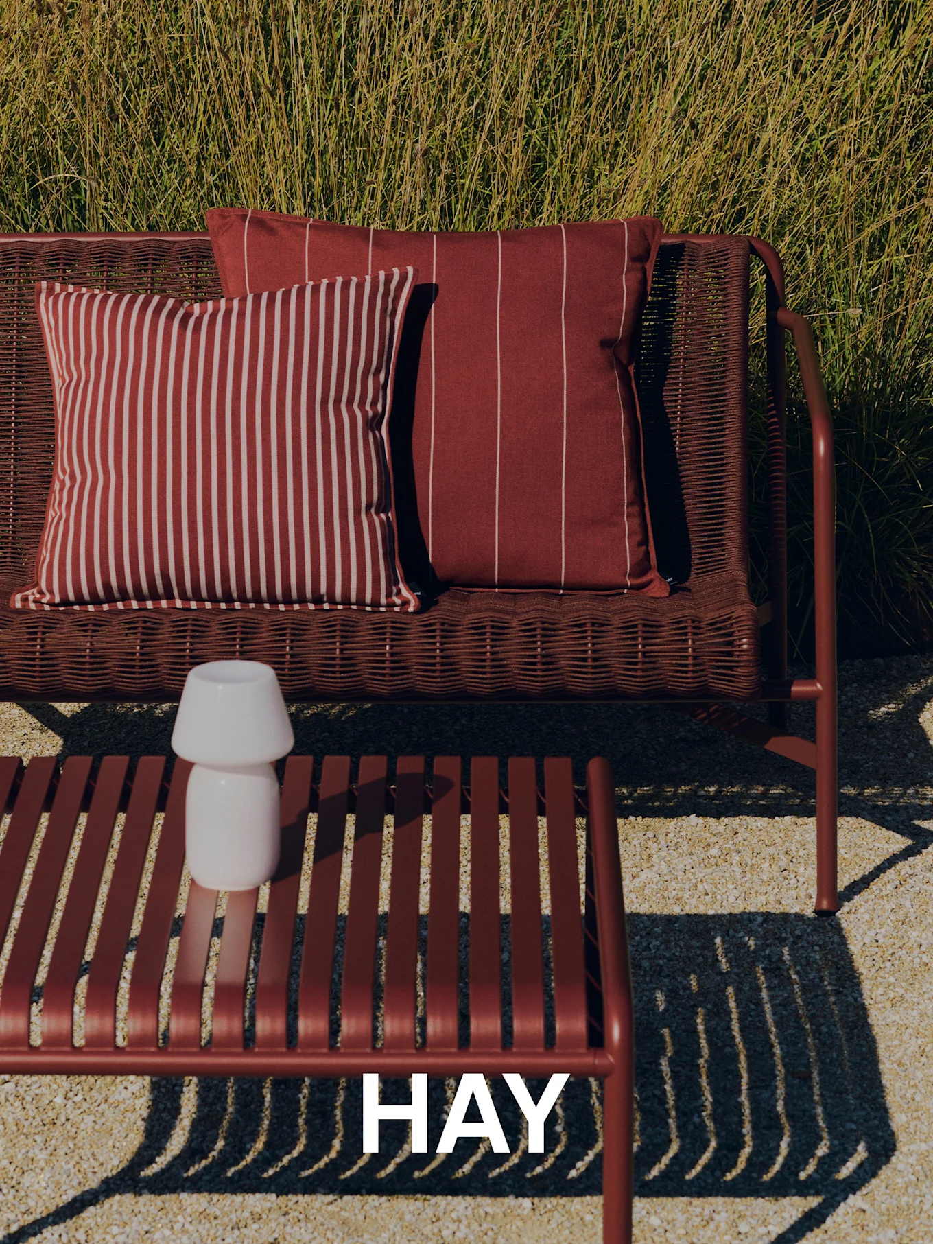 Scena zewnętrzna z plecioną czerwoną sofą, poduszkami w paski, stolikiem kawowym z listew z białą lampą i wysoką trawą w tle. Logo HAY.