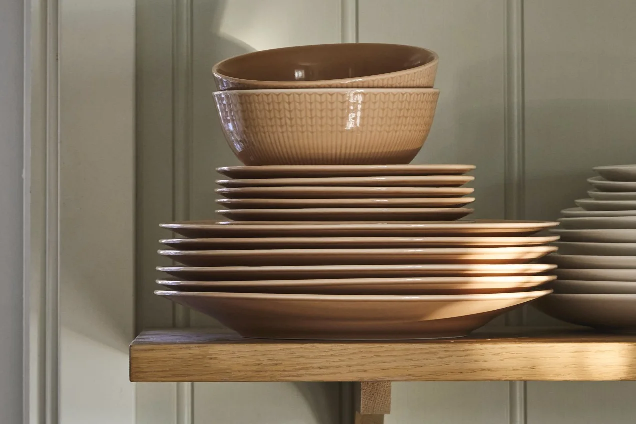 Stosy jasnobrązowych ceramicznych talerzy i wzorzystych misek na drewnianej półce.