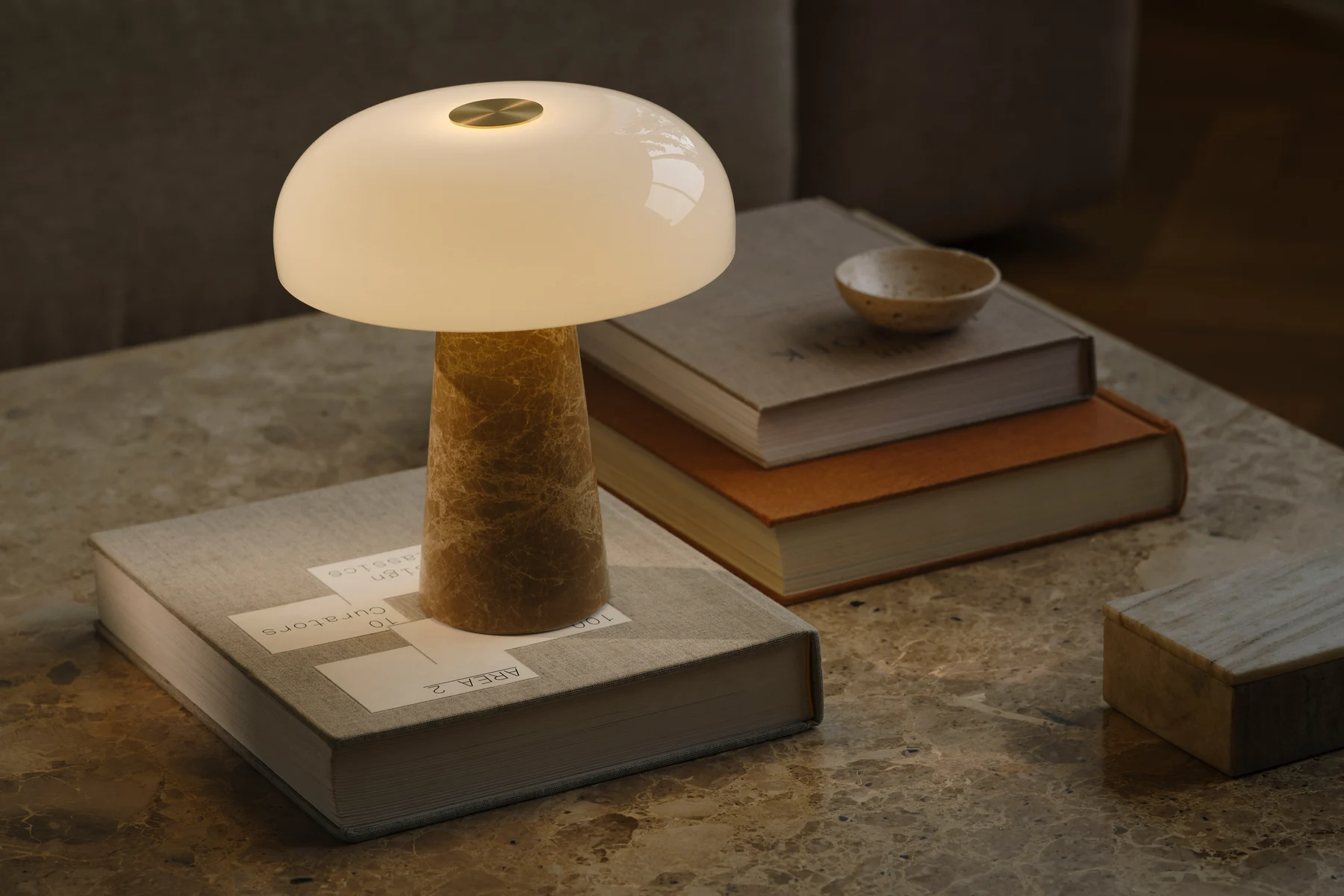 Lampa stołowa w kształcie grzyba z białym szklanym kloszem i stożkową marmurową podstawą stoi na marmurowym stole z ułożonymi książkami.