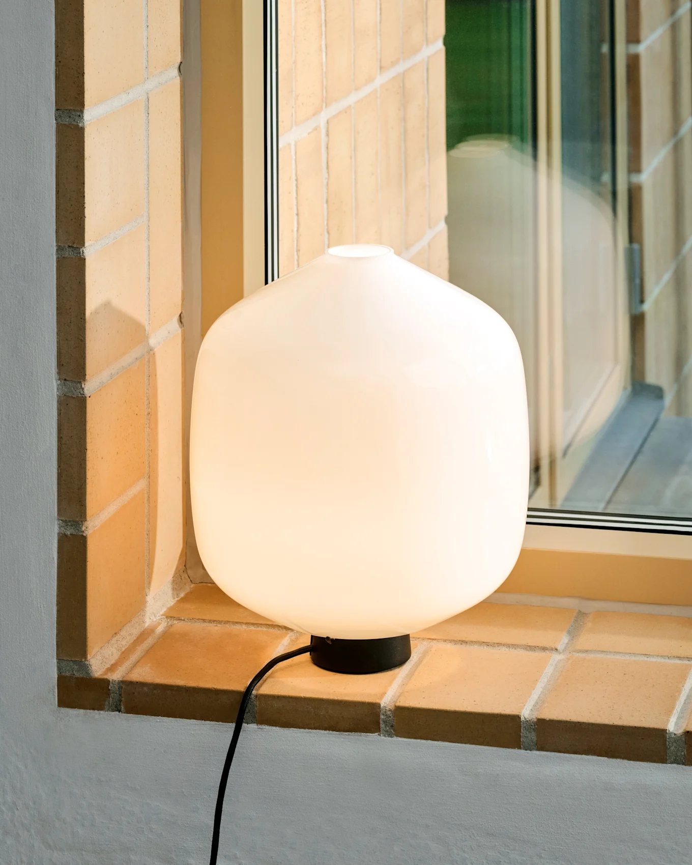 Świecąca biała zaokrąglona lampa stołowa z czarną podstawą i przewodem stoi na jasnym ceglanym parapecie.