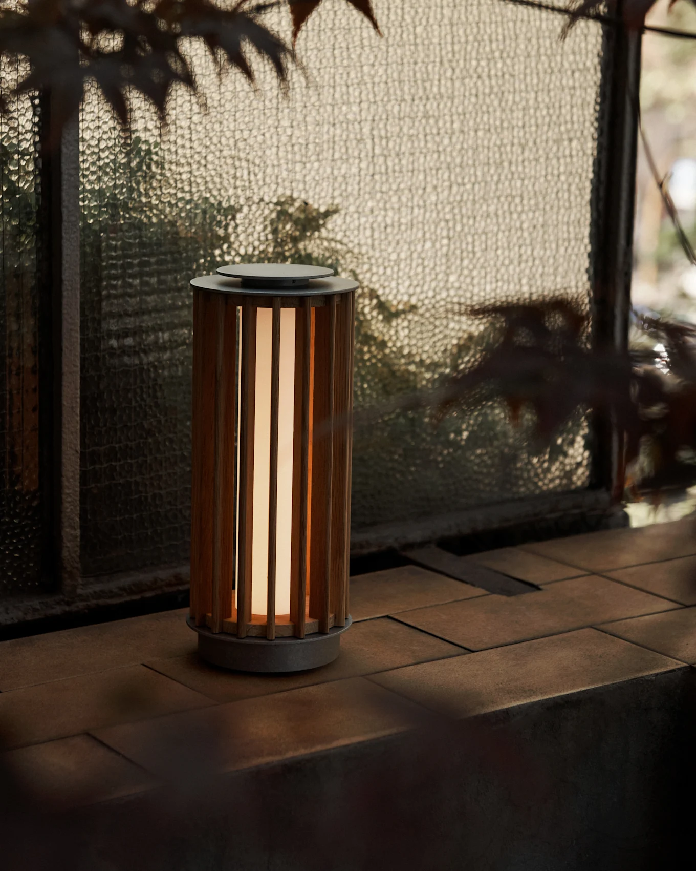 Cylindryczna lampa przenośna z drewnianą obudową z listew i ciepłym blaskiem, stojąca na kafelkowej powierzchni przy teksturowanym szklanym oknie.