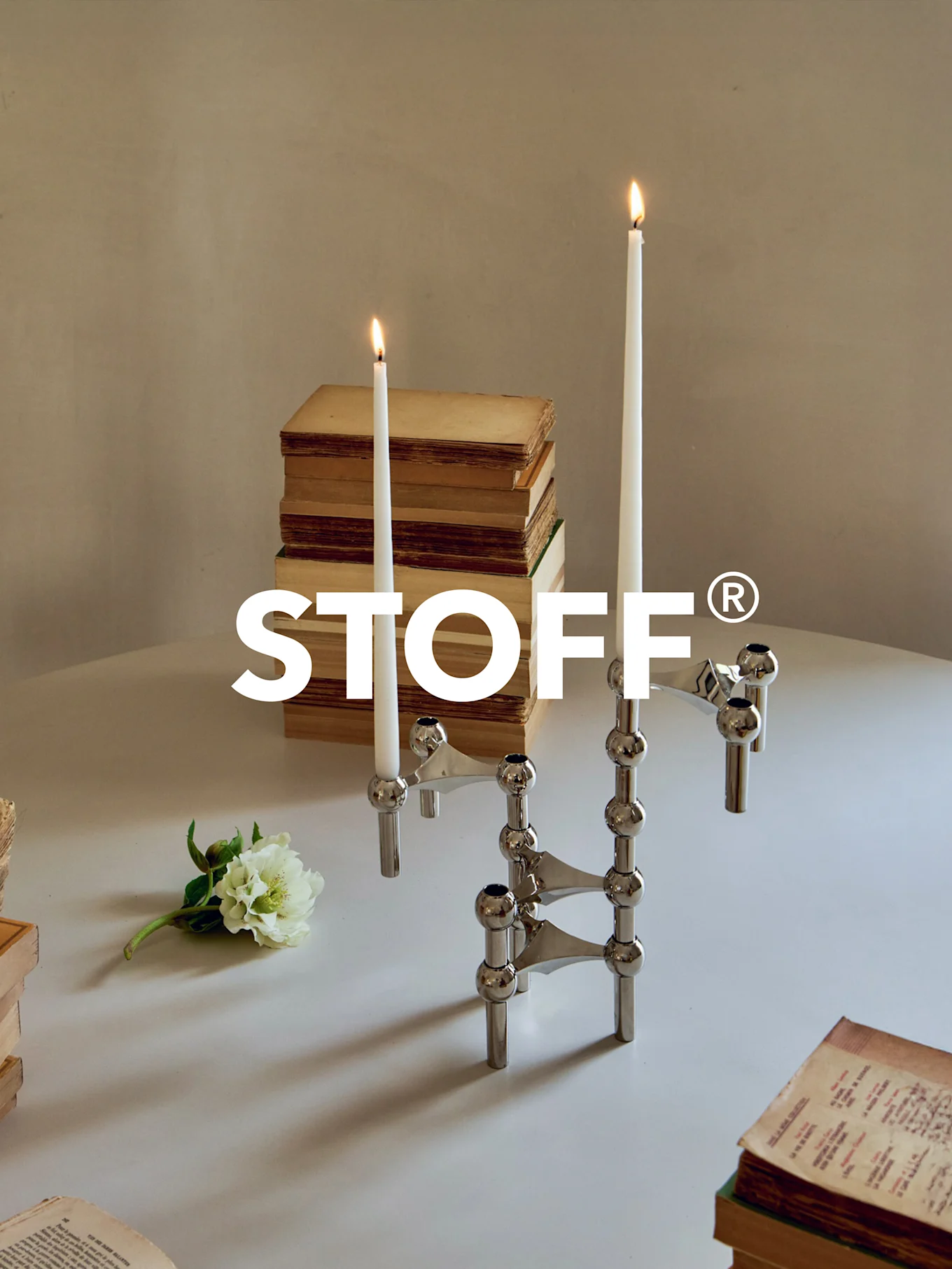 Polerowany chromowany modułowy świecznik z dwoma zapalonymi świecami, otoczony starymi książkami i białym kwiatem na stole. Logo STOFF®.