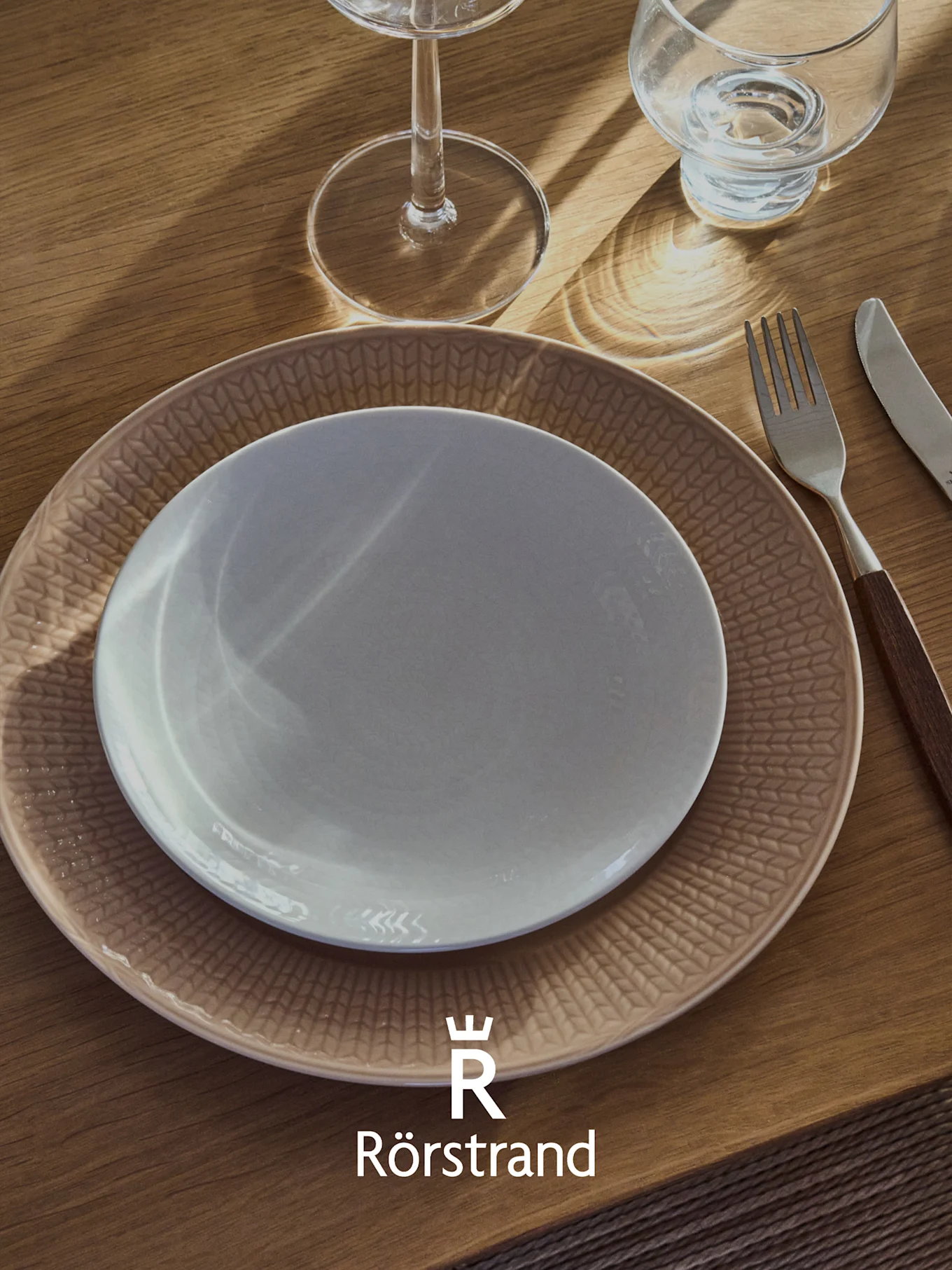 Nakrycie stołu z białym talerzem na beżowym talerzu we wzory, widelcem, nożem i dwoma szklankami na drewnianym stole. Logo Rörstrand.