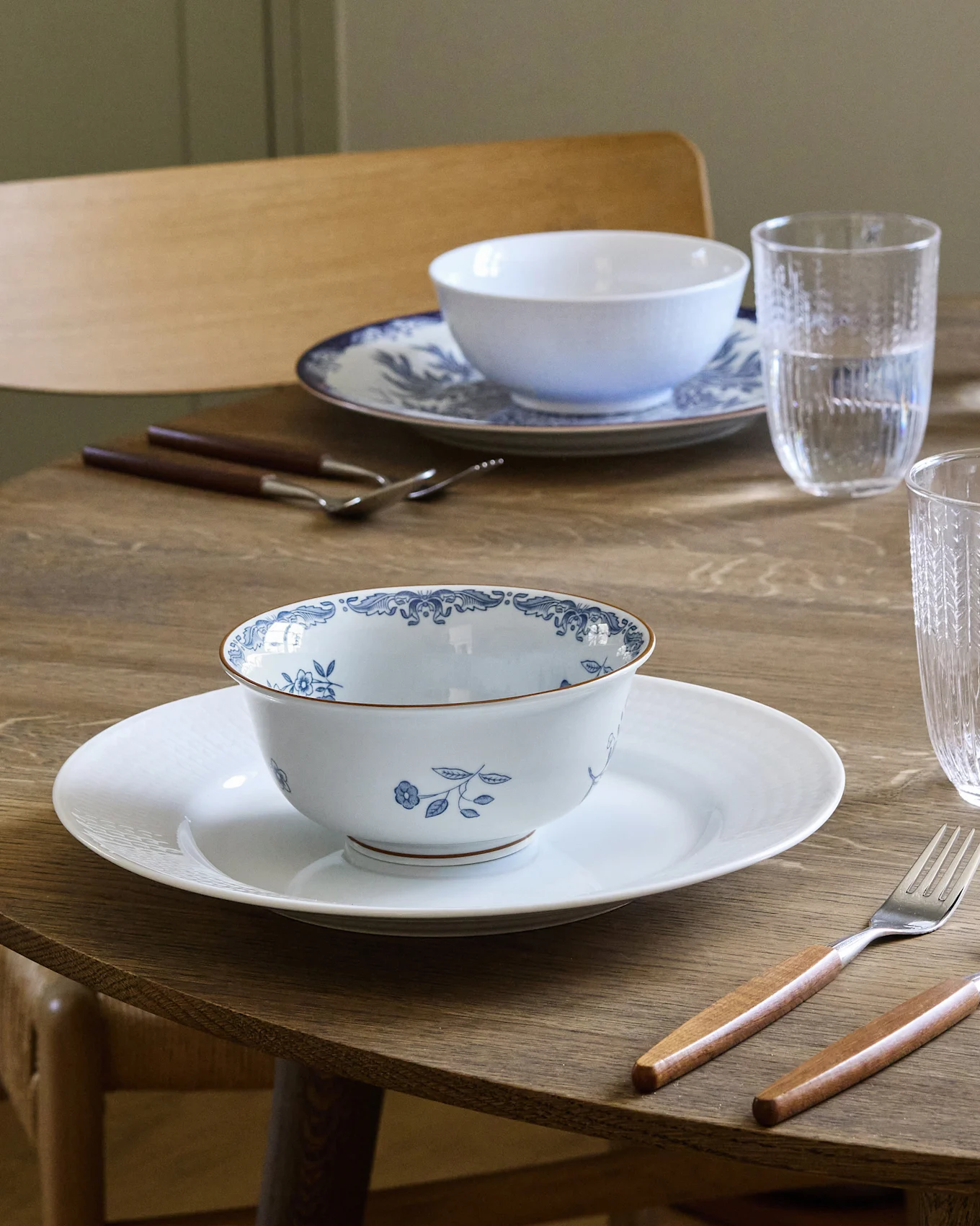 Stół jadalny nakryty dla dwóch osób z białymi i niebieskimi miskami z wzorami na białych talerzach, sztućcami z drewnianymi uchwytami i szklankami na wodę.