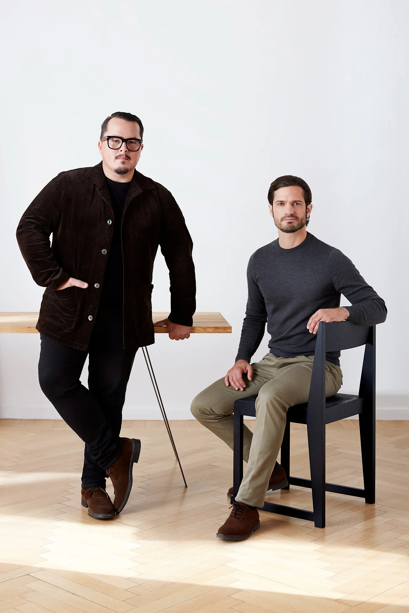 Dwóch mężczyzn, jeden stoi przy drewnianym stole w brązowej sztruksowej kurtce, drugi siedzi na czarnym krześle w szarej koszuli.