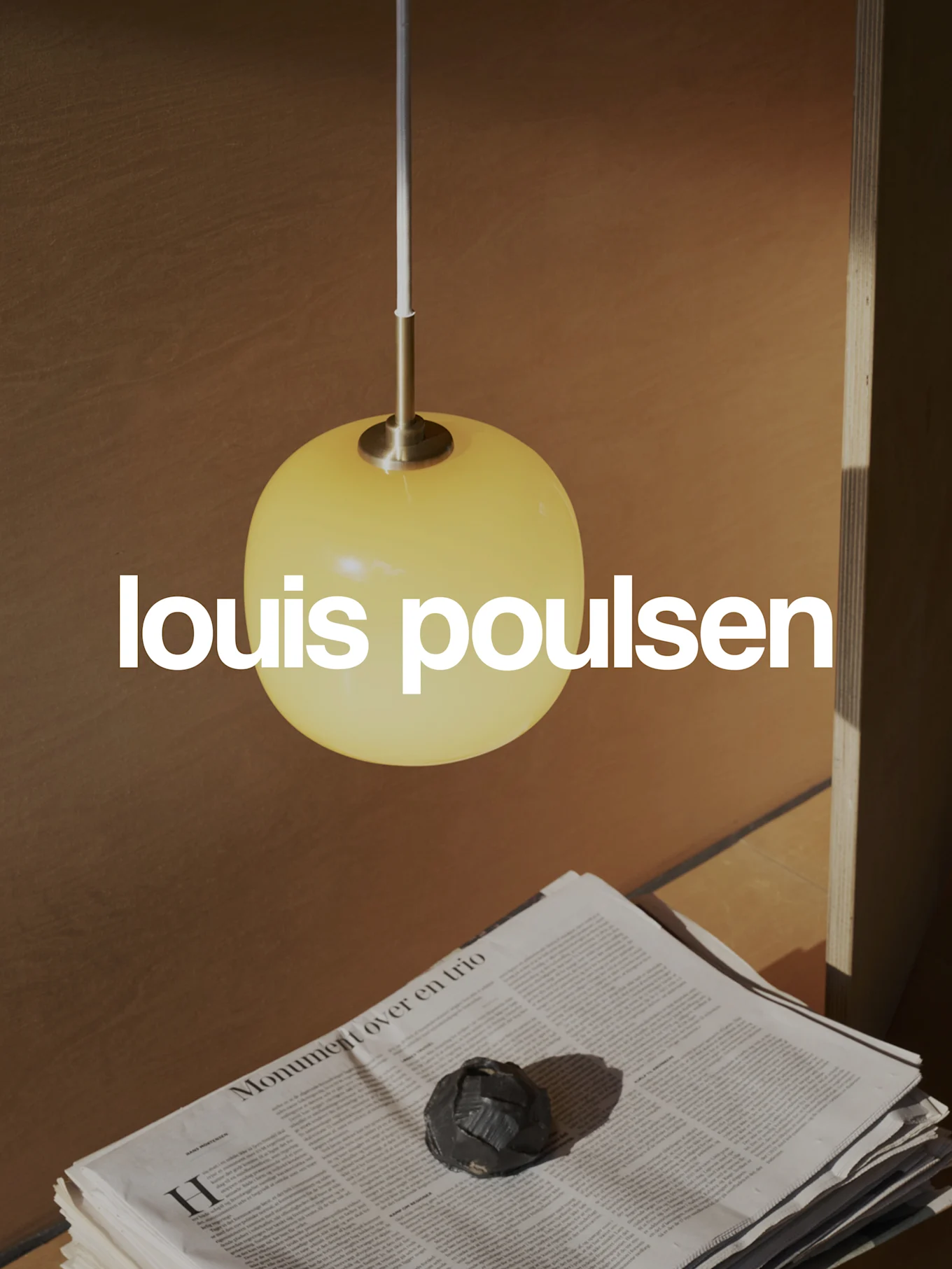 Logo Louis Poulsen na tle ciepłej sceny z żółtą kulistą lampą wiszącą, stosami gazet i ciemnym przedmiotem dekoracyjnym.