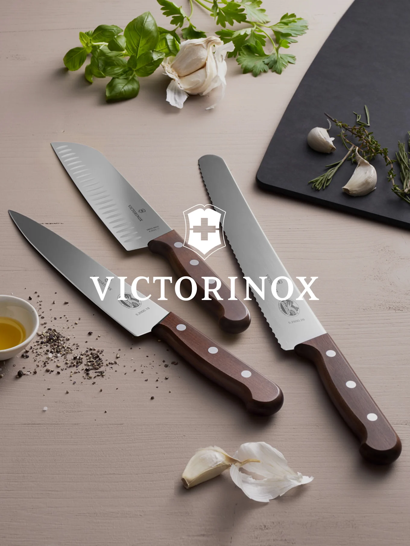 Trzy noże Victorinox z drewnianymi rączkami, świeże zioła i czosnek na drewnianej powierzchni.