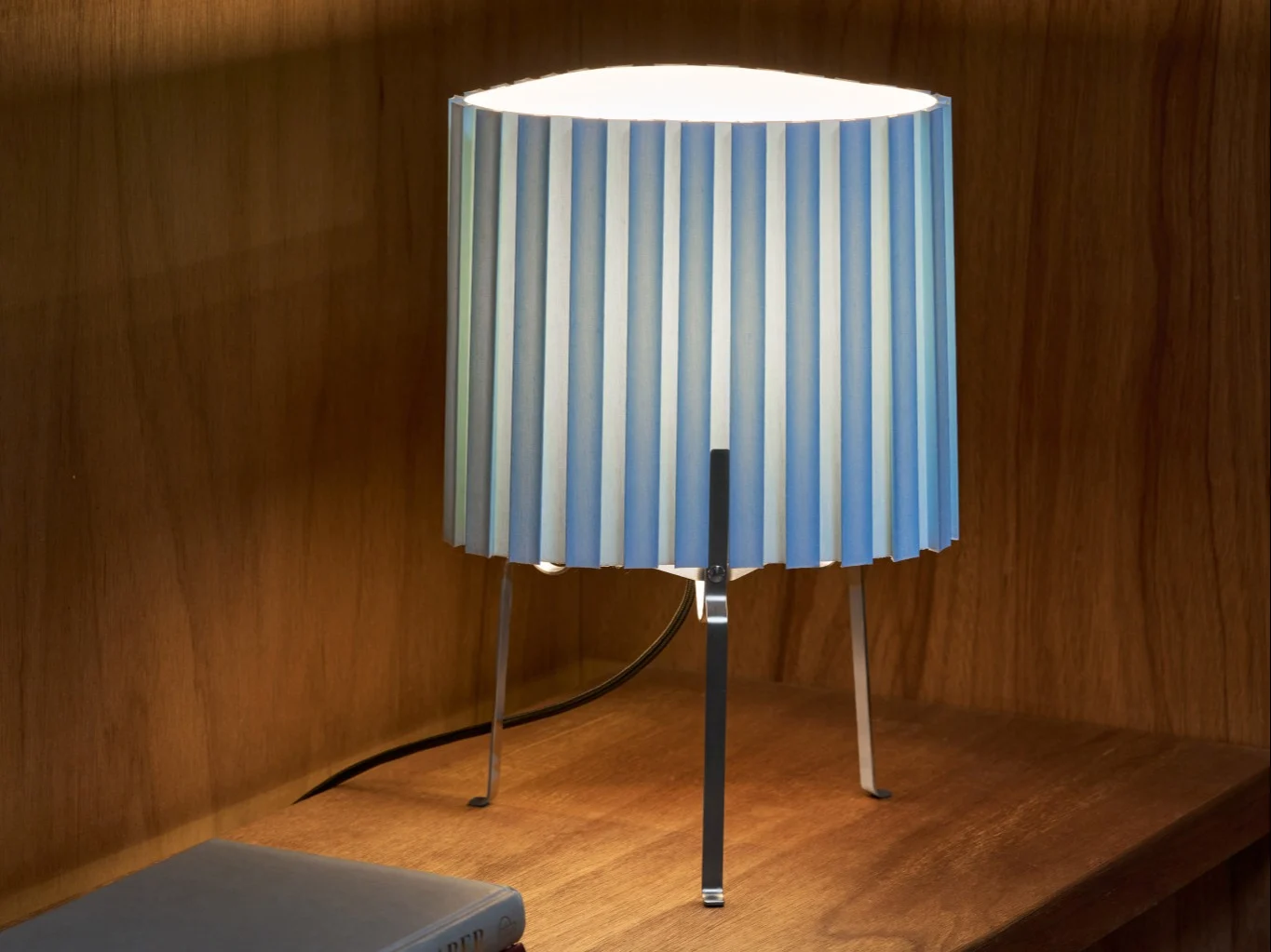 Lampa stołowa z plisowanym abażurem w niebiesko-białe pasy na drewnianej półce, oświetlająca otoczenie.