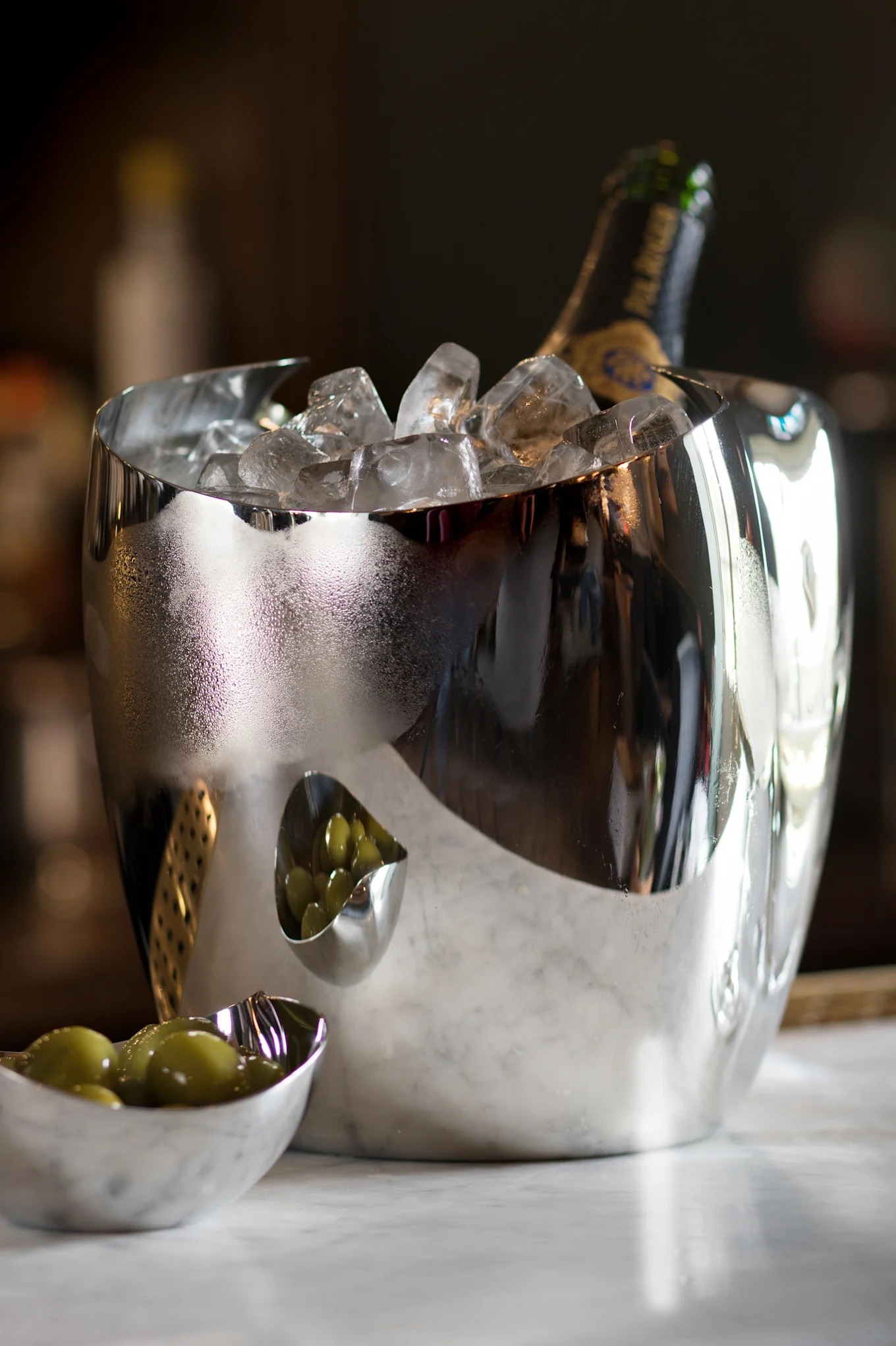 Odblaskowe srebrne wiaderko na lód z butelką szampana i lodem, obok dwóch małych miseczek z zielonymi oliwkami na marmurze.