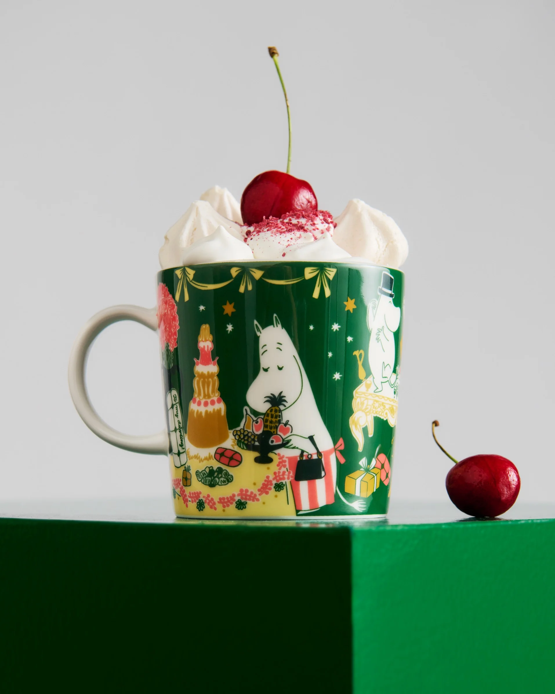Ciemnozielony kubek z postaciami Moomin, zwieńczony bitą śmietaną, czerwoną posypką i wiśnią. Kolejna wiśnia leży na zielonej powierzchni.