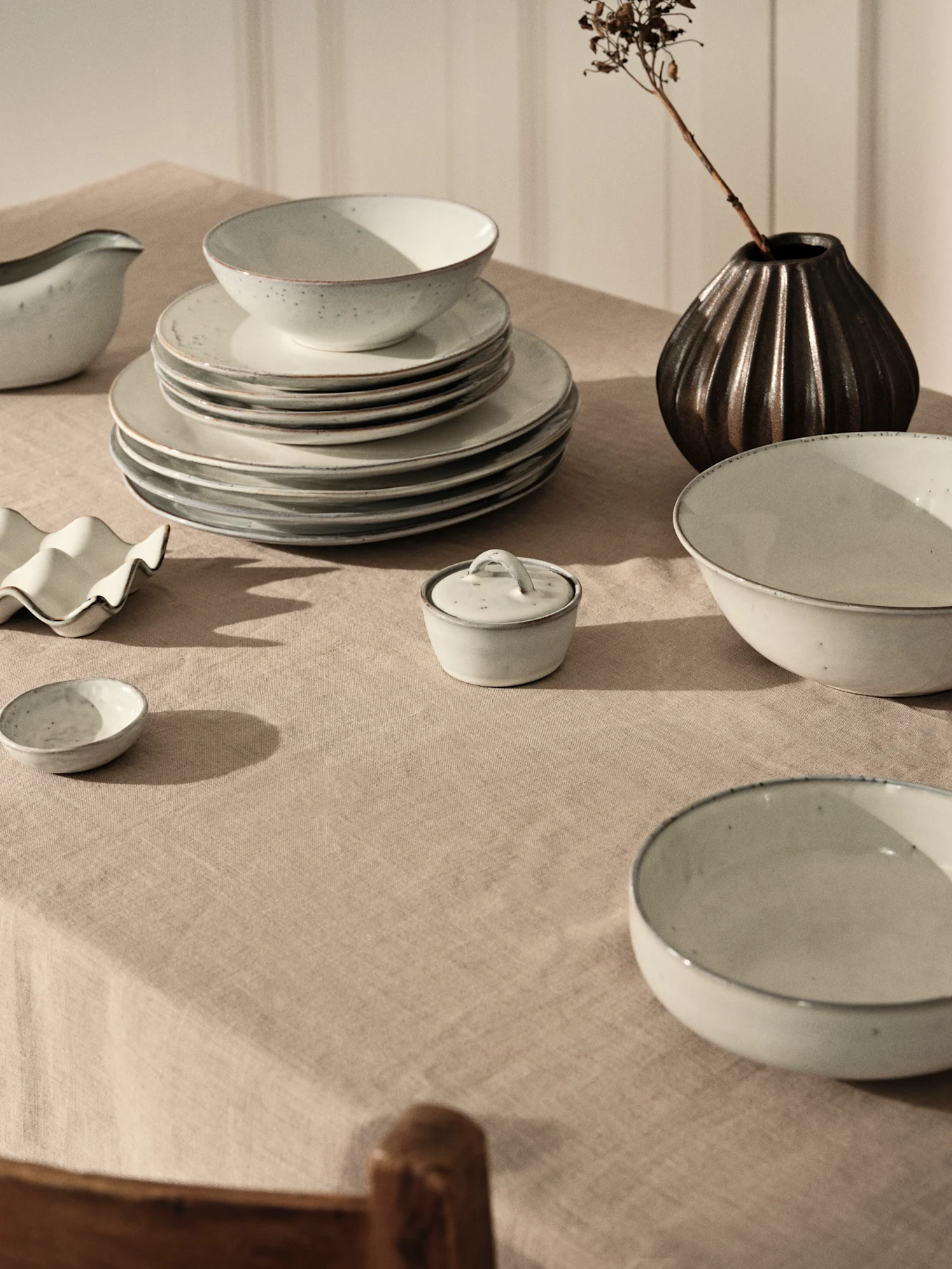 Jasnoszary ceramiczny zestaw, stosy talerzy i misek na beżowym lnianym obrusie, obok ciemna waza z suszoną gałęzią.