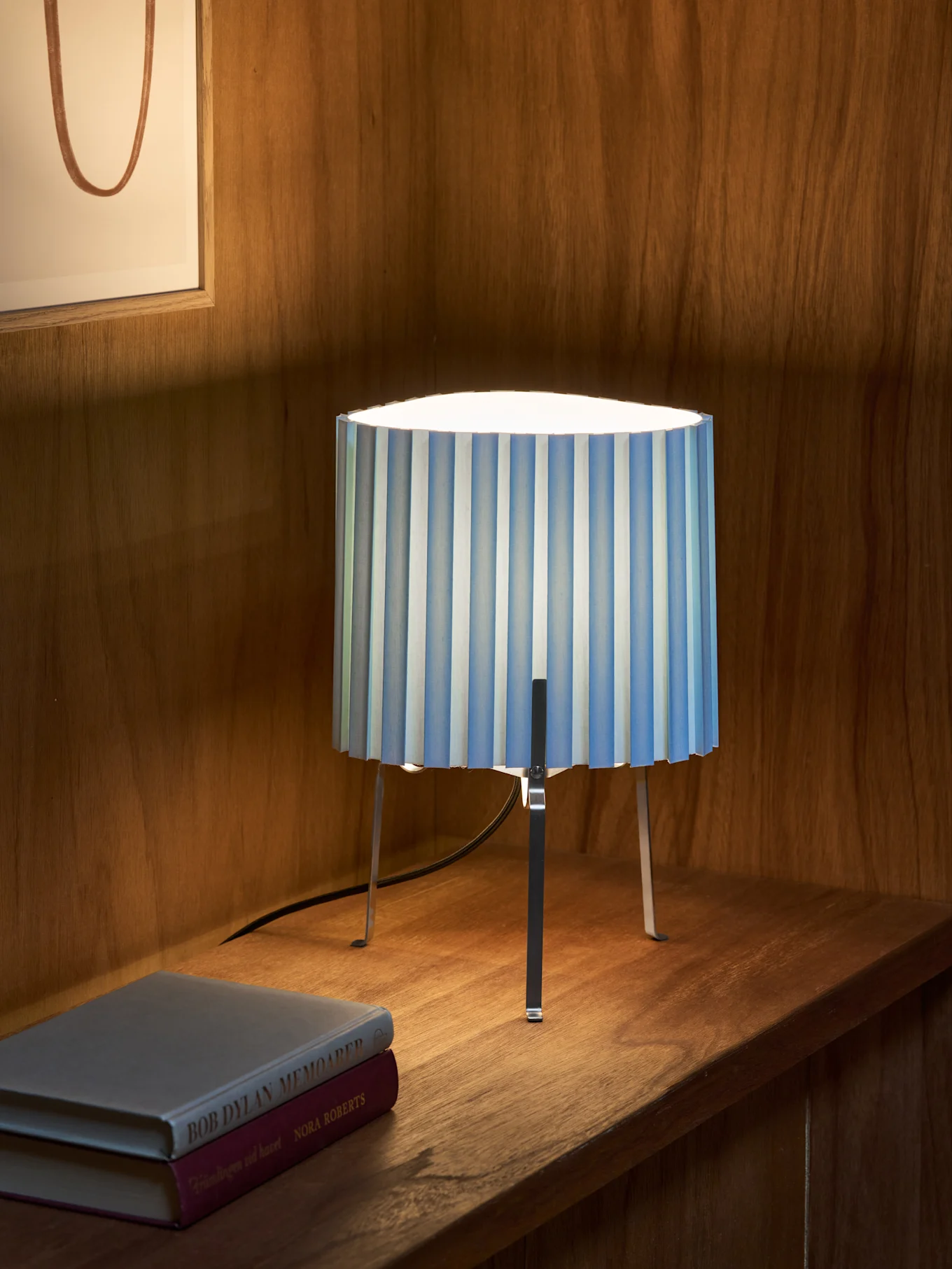 Lampa stołowa w niebiesko-białe pasy z trzema metalowymi nogami stoi na drewnianej półce obok dwóch książek. W tle oprawiona grafika.