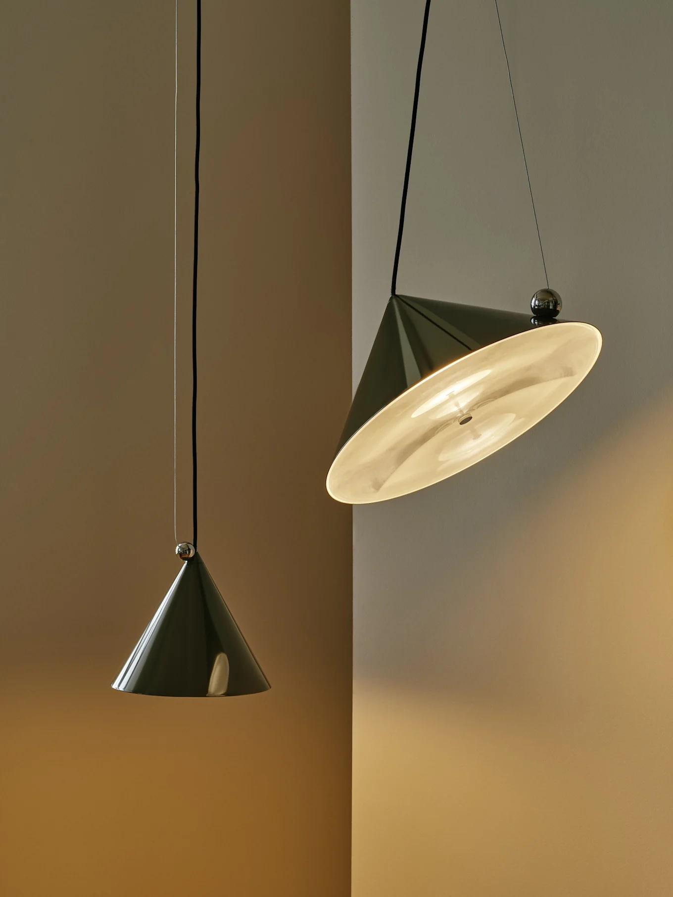 Dwie ciemnozielone stożkowe lampy wiszące na dwukolorowej ścianie; jedna jest zapalona i pochylona, druga wisi pionowo.