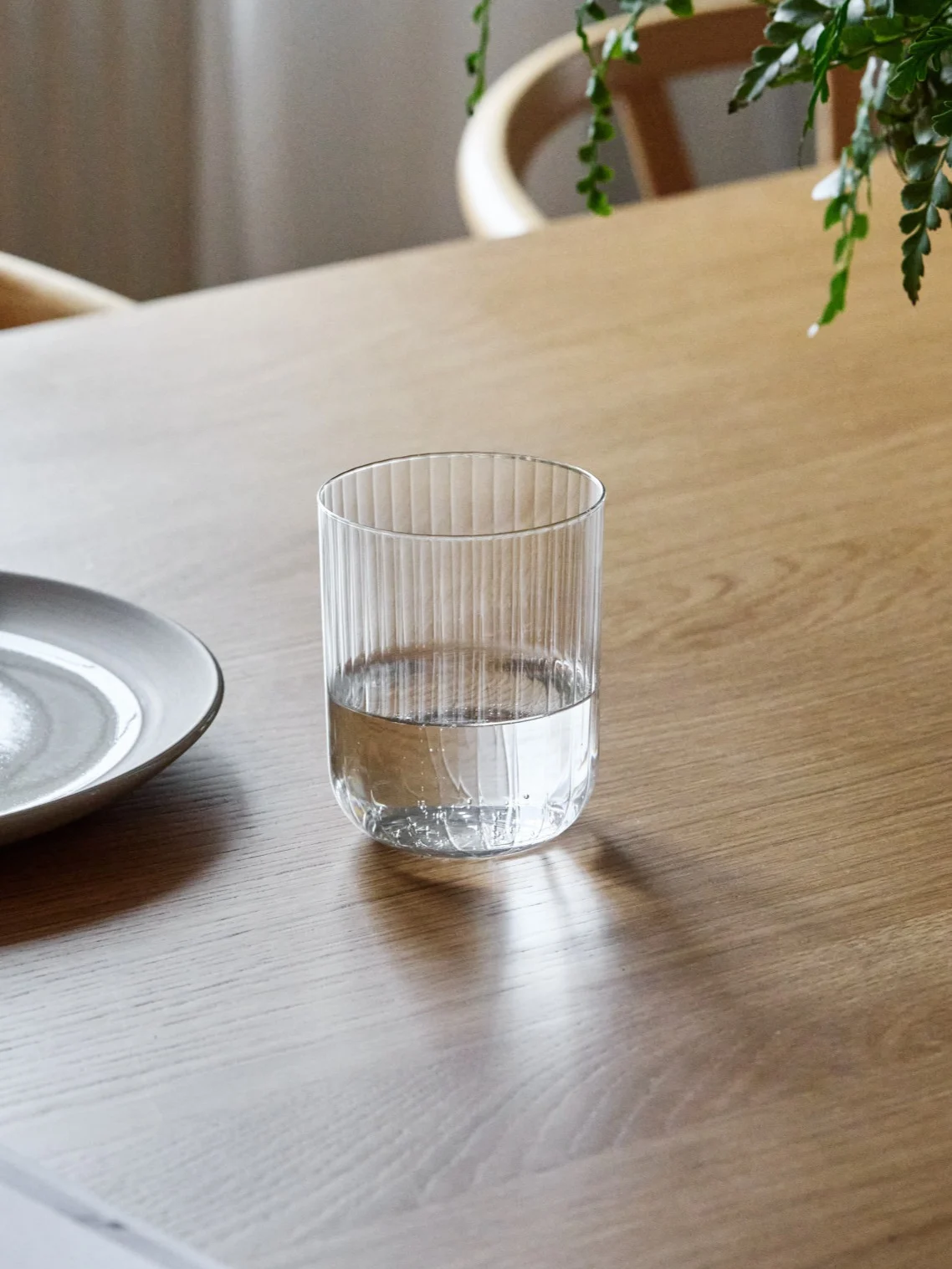 Żłobkowana szklanka, w połowie wypełniona wodą, stoi na drewnianym stole obok ceramicznego talerza.