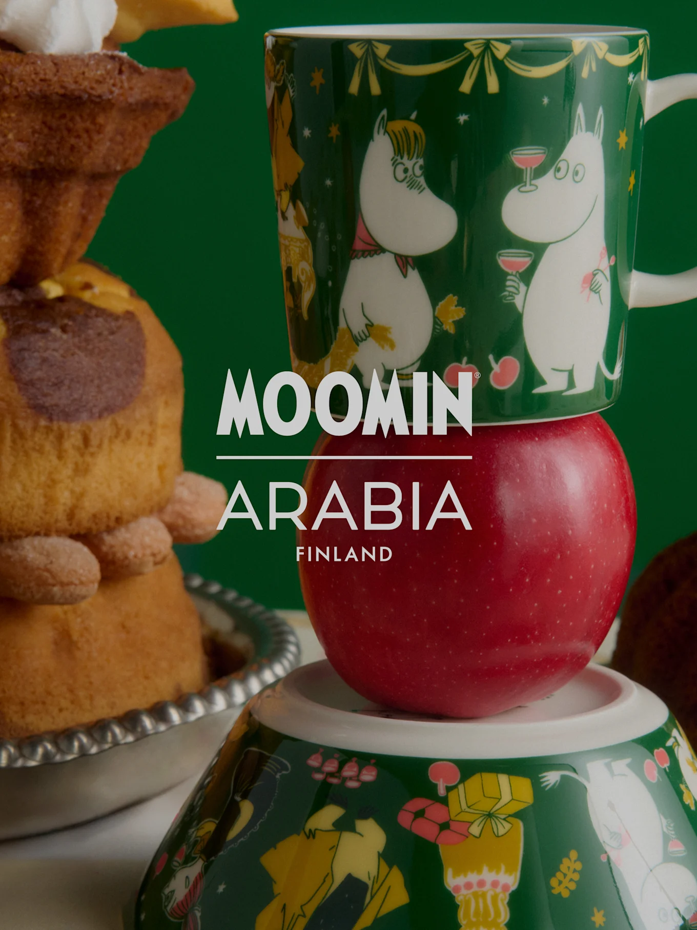 Świąteczna kolekcja Moomin Arabia: Zielony kubek Moomin na czerwonym jabłku, na misce Moomin, obok świątecznych wypieków.