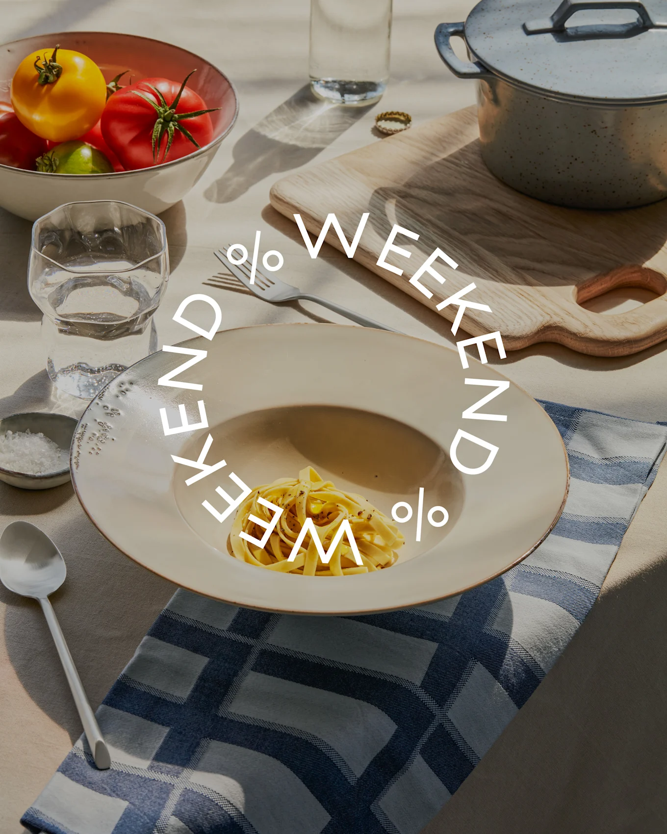 Stół z makaronem, miską świeżych pomidorów, szklanką wody i garnkiem. Tekst 'WEEKEND % WEEKEND %' jest nałożony.