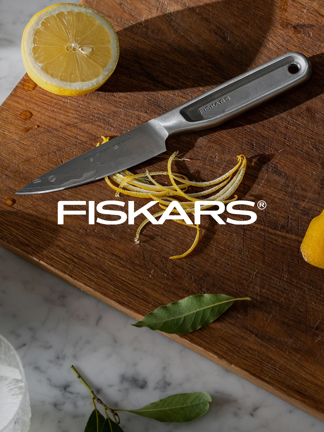 Nóż Fiskars na drewnianej desce do krojenia z połówką cytryny, skórką cytrynową i kroplami wody.