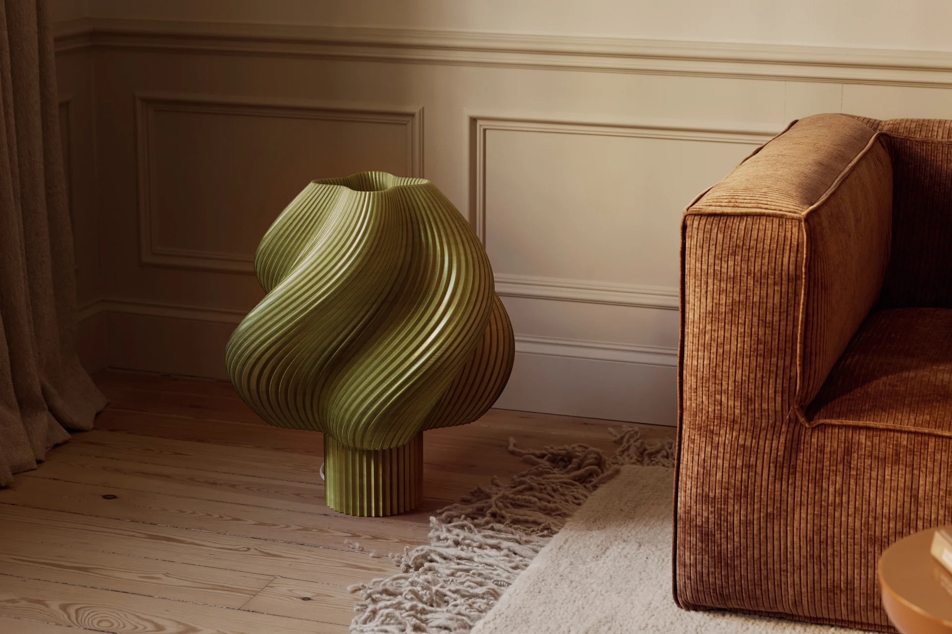 Oliwkowozielony, rzeźbiarski, żłobiony obiekt na drewnianej podłodze obok brązowej sztruksowej sofy i teksturowanego dywanu w pokoju z formowanymi ścianami.