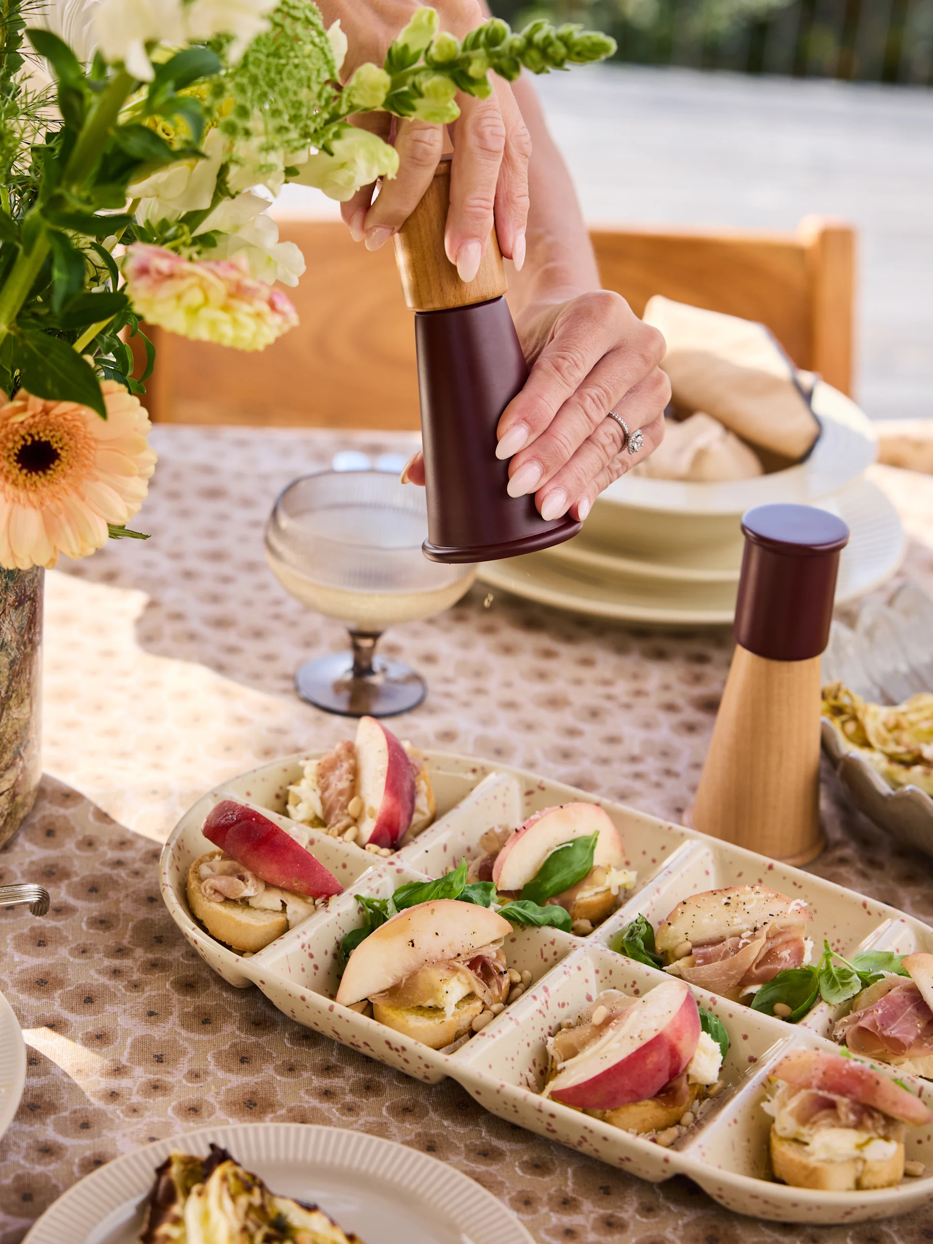 Dłonie mielące pieprz na crostini z brzoskwiniami, prosciutto, serem, orzeszkami piniowymi i bazylią.