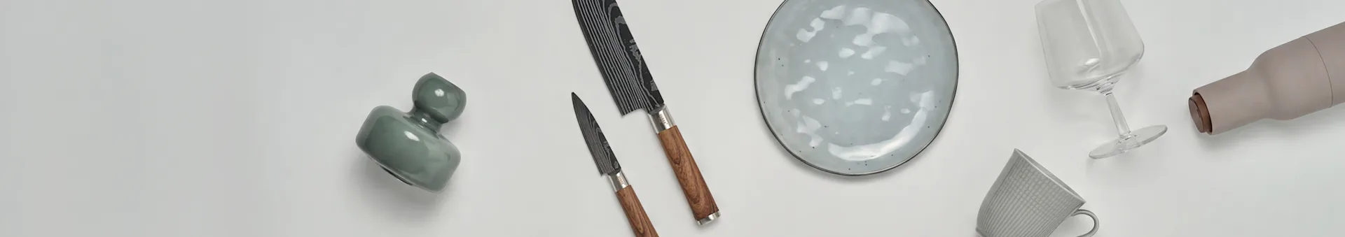 Różne artykuły kuchenne i jadalniane, w tym zielony wazon, dwa noże, nakrapiany talerz, kieliszek do wina, beżowa butelka i wzorzysty kubek.