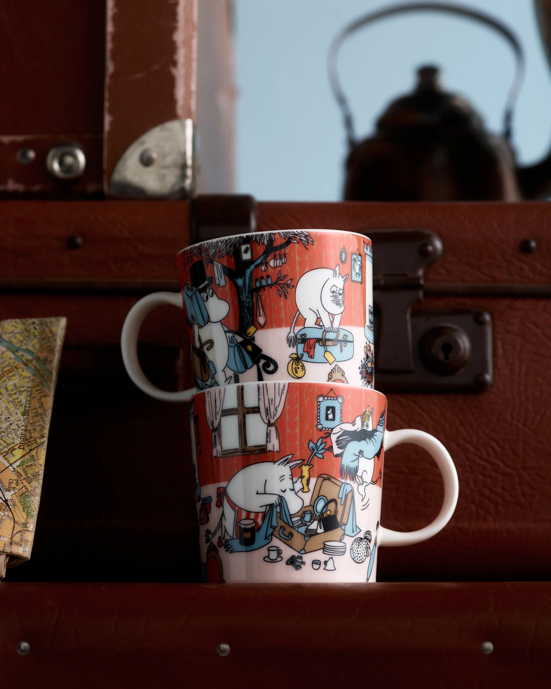 Dwa kubki Moomin z pomarańczowo-czerwonymi ilustracjami postaci, ułożone jeden na drugim przed brązowymi walizkami i niewyraźną mapą.