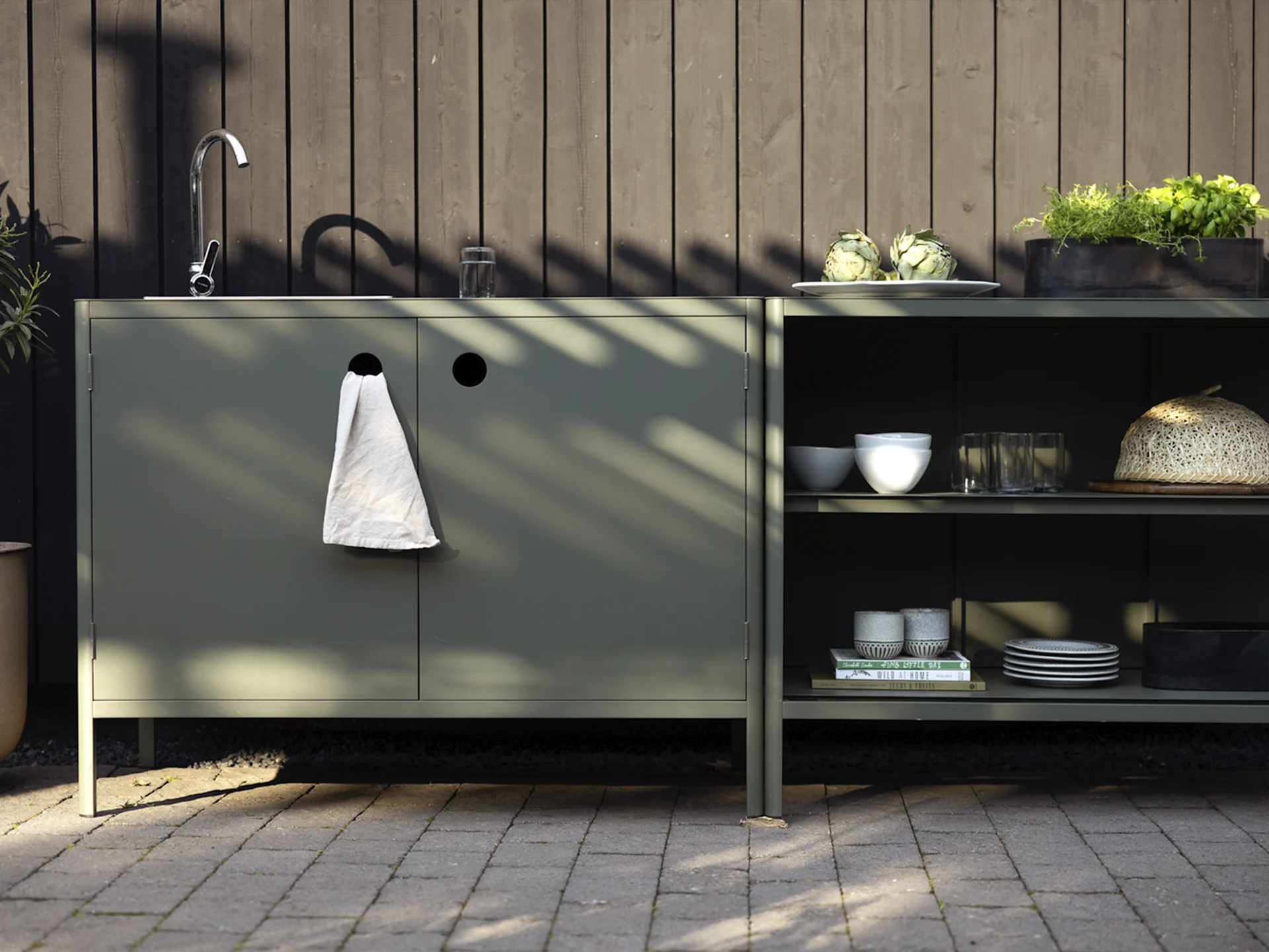 Kuchnia zewnętrzna z ciemnozielonymi szafkami, zlewem, kranem i półkami z karczochami, ziołami i zastawą.