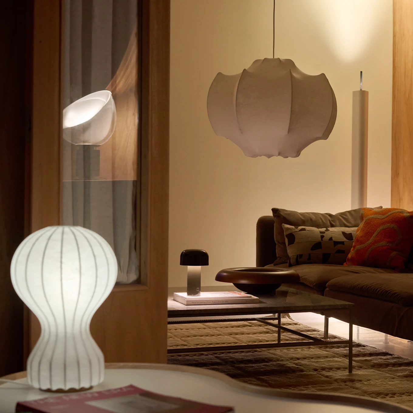 Salon z brązową sofą i stolikiem kawowym, oświetlony białą żeberkową lampą stołową, ciemną lampą grzybową i dużą plisowaną lampą wiszącą, tworząc ciepły blask.