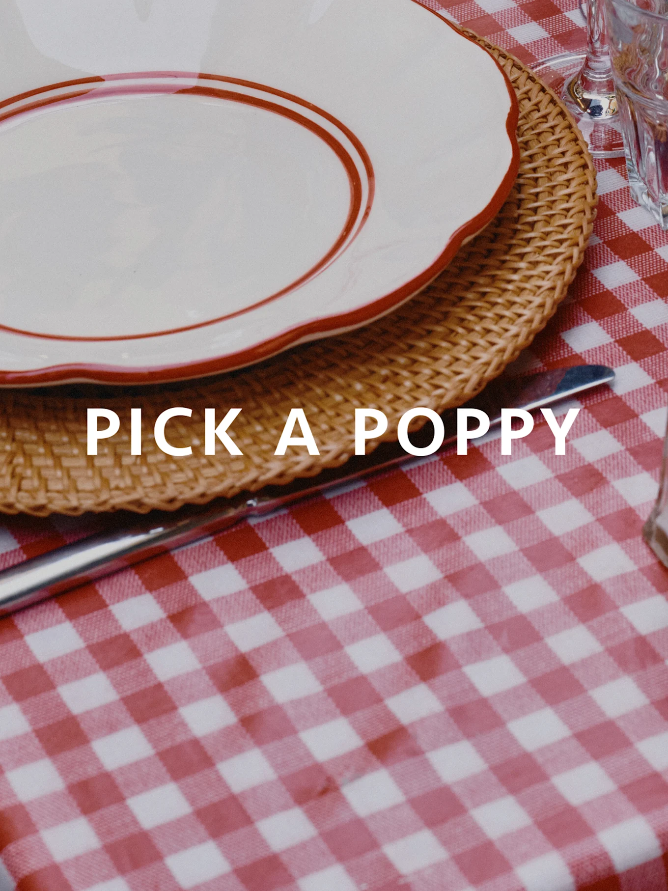 Nakryty stół z czerwoną i białą obruszką w kratkę, wiklinową podkładką i białym talerzem z czerwoną obwódką. Tekst: PICK A POPPY.