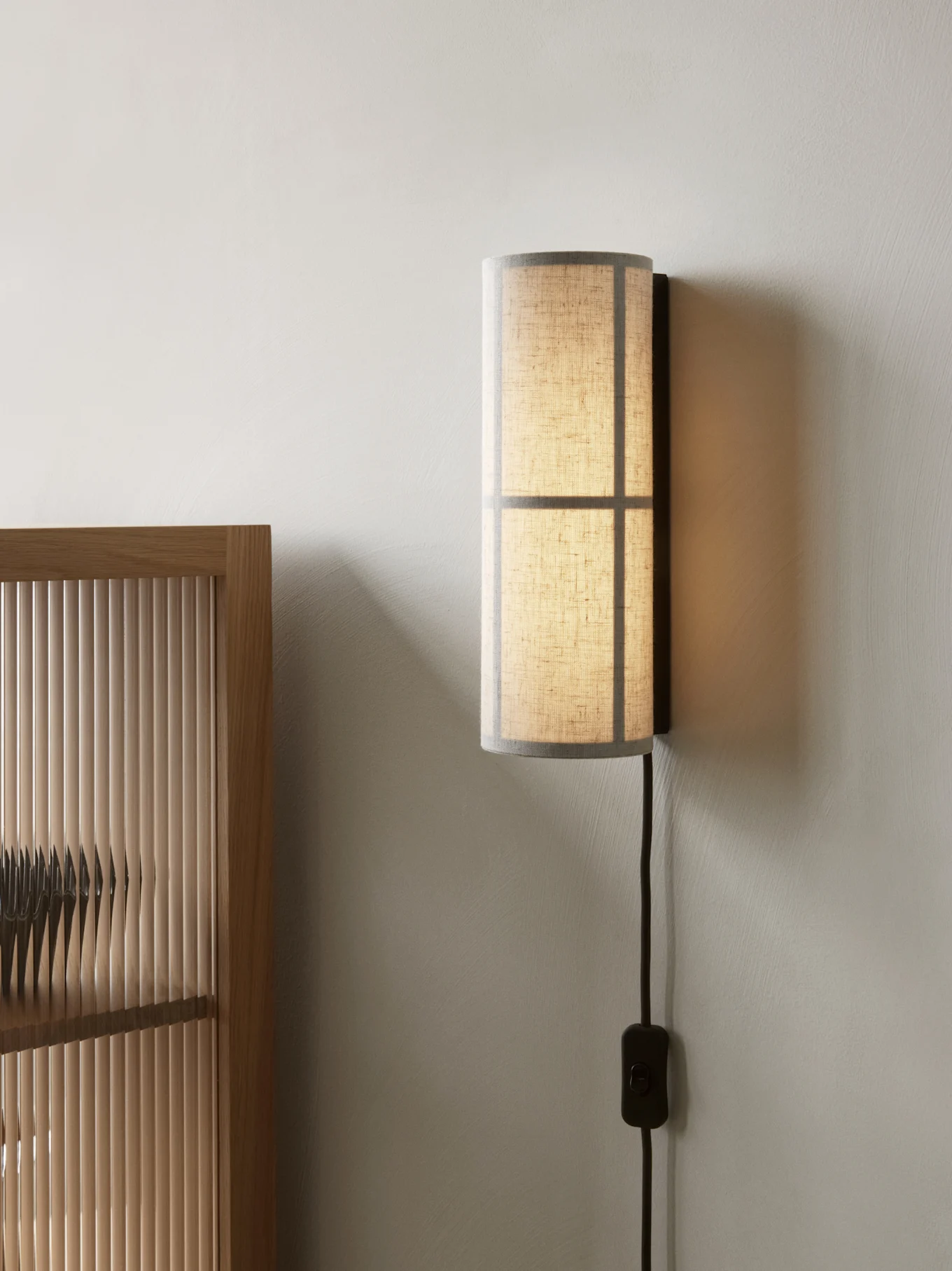 Cylindryczna lampa ścienna z przezroczystym abażurem z tkaniny, zamontowana na jasnej ścianie obok drewnianej ścianki.