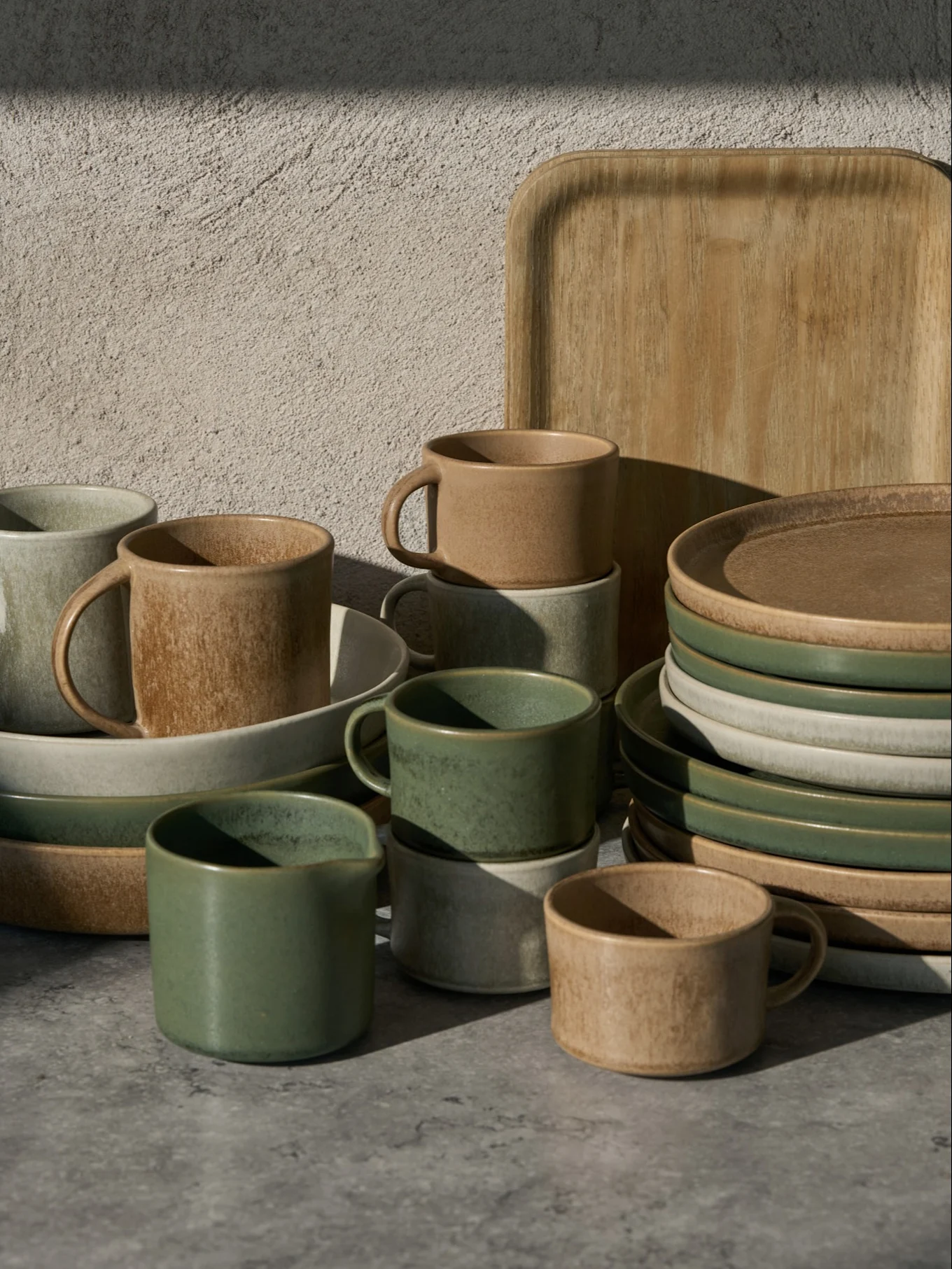 Kolekcja zielonych, beżowych i jasnoszarych ceramicznych filiżanek, misek i talerzy, z drewnianą tacą, na szarej powierzchni.