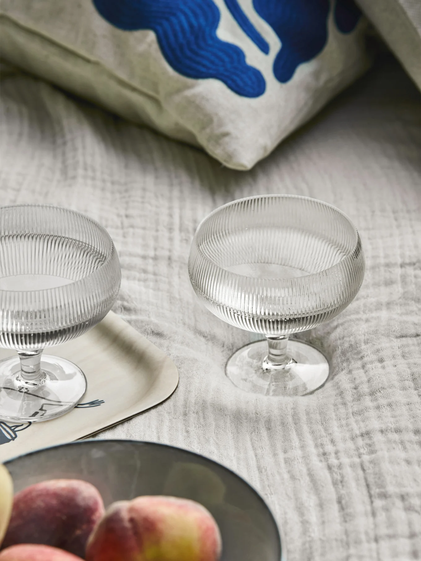 Dwie szklane miseczki na deser z żebrowanym wzorem, poduszka z niebieską sztuką abstrakcyjną, drewniana taca i brzoskwinie na jasnej tkaninie.