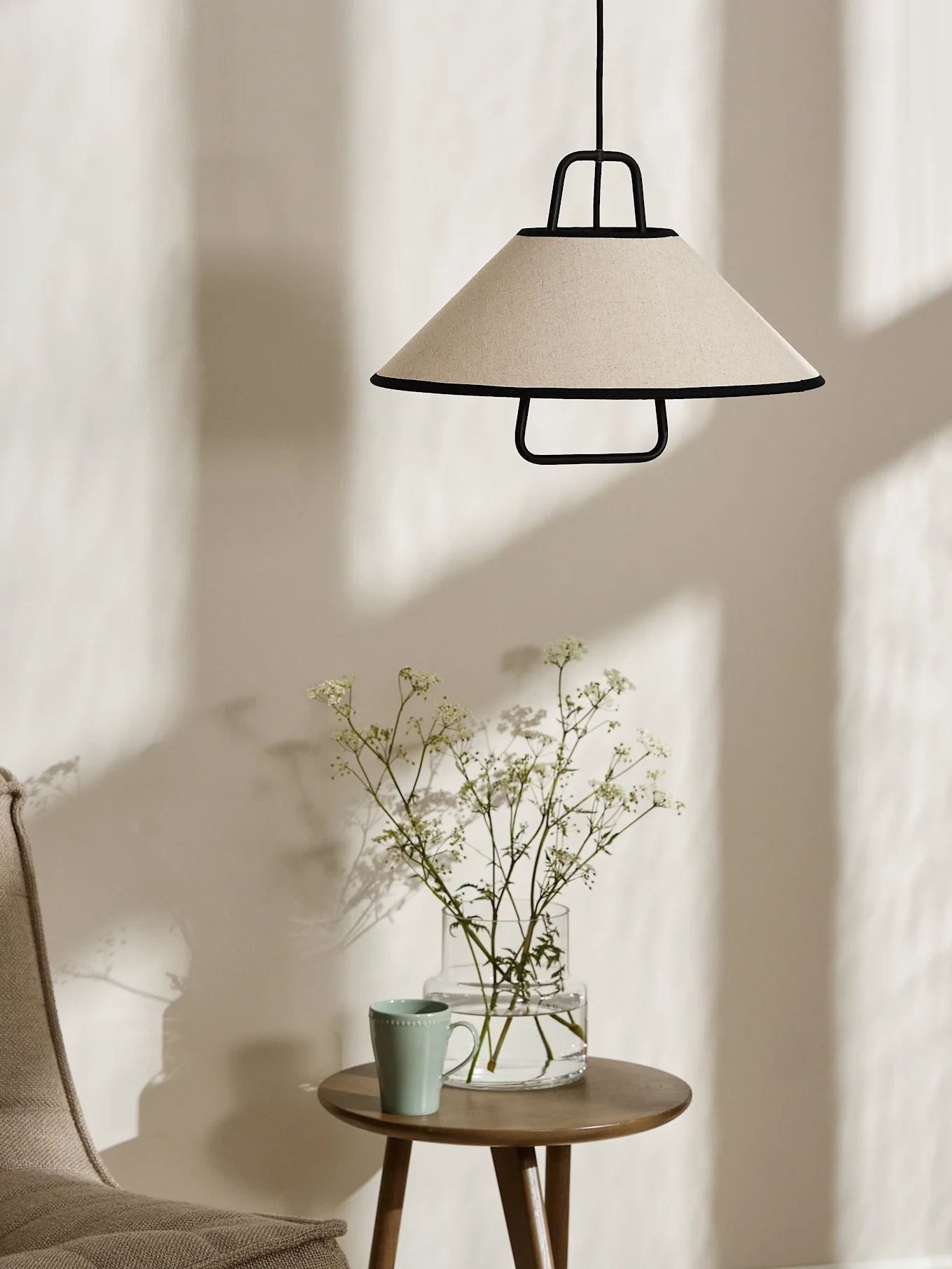 Stożkowa lampa wisząca w kolorze kremowym z czarnym obramowaniem wisi nad drewnianym stolikiem bocznym z wazonem białych kwiatów i jasnozielonym kubkiem.