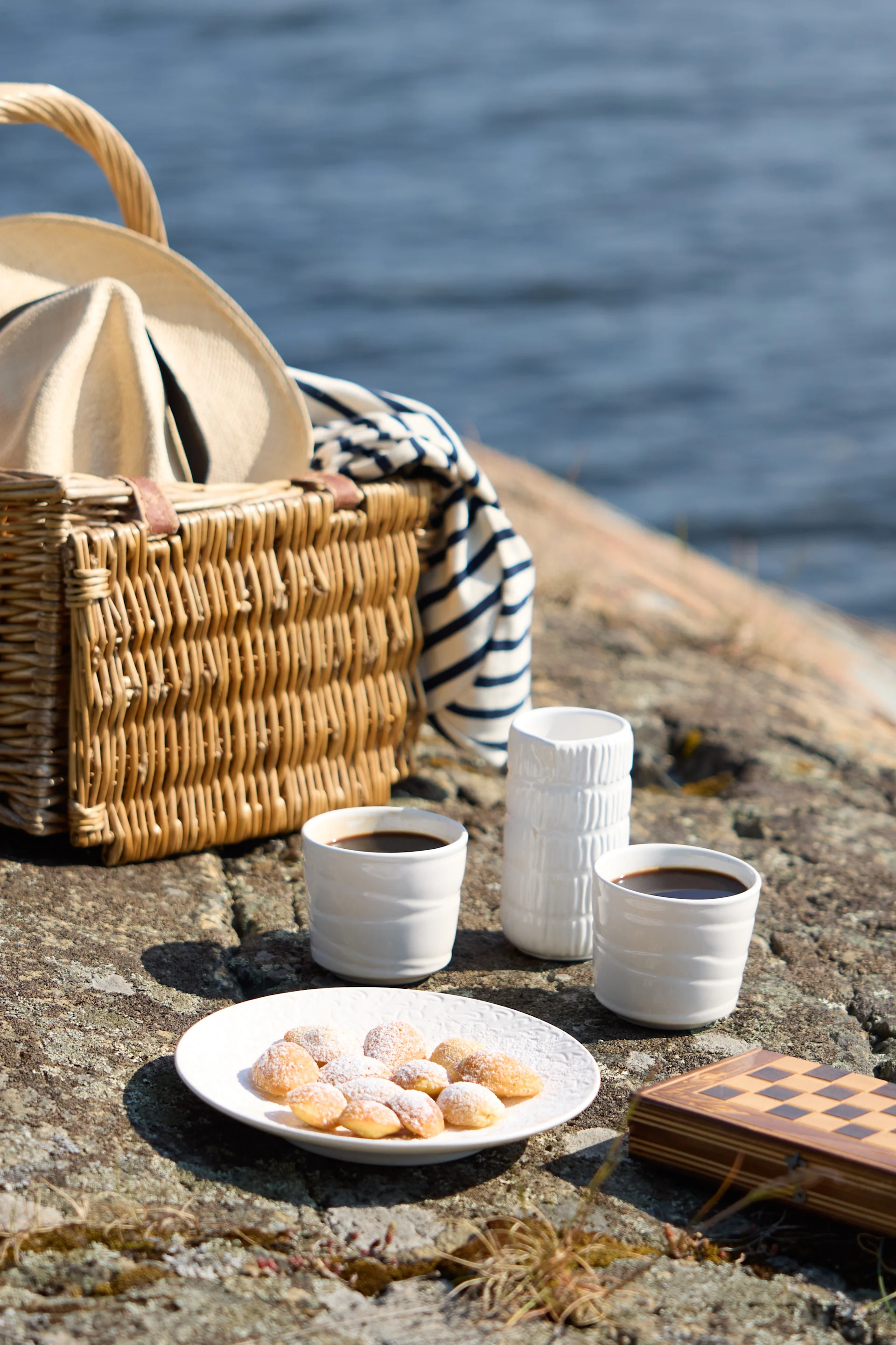 Piknik na skałach nad morzem, z wiklinowym koszem z kapeluszem i kocem w paski, dwoma filiżankami kawy, ciastkami i zamkniętą szachownicą.