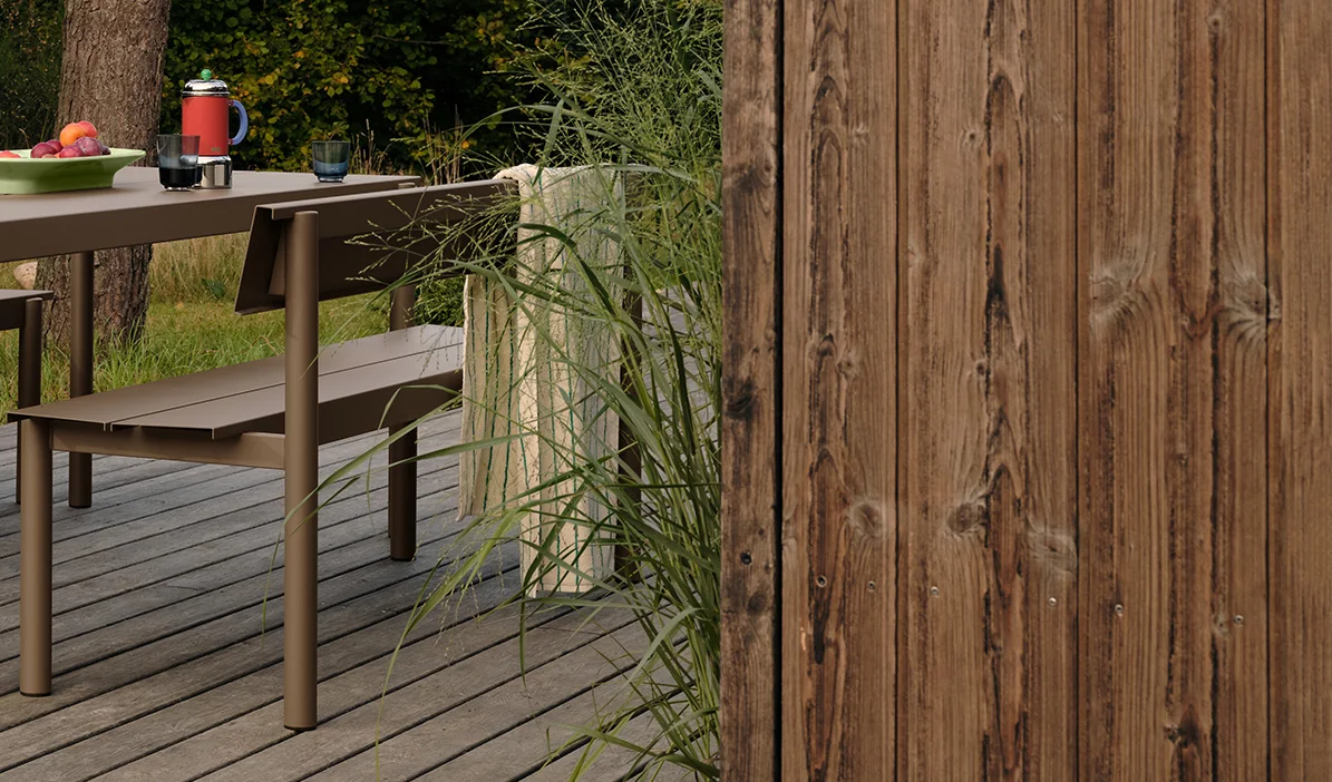 Brązowy metalowy stół i ławka ogrodowa na drewnianym tarasie z owocami i napojami, obok wysokiej trawy i drewnianego płotu.