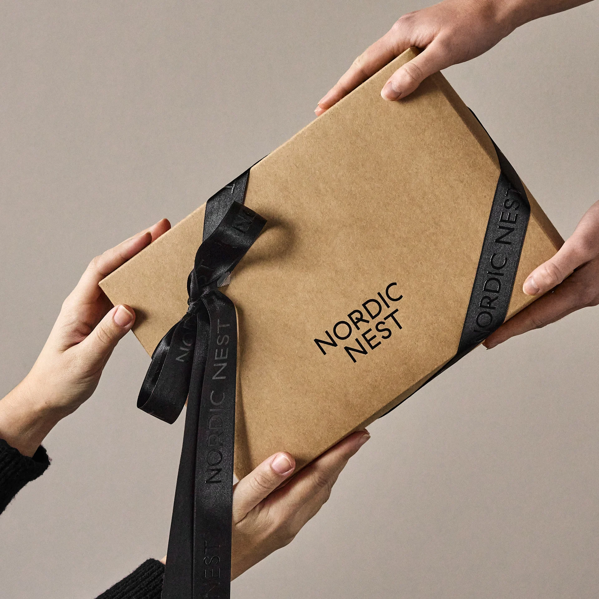 Dłonie wymieniające brązowe pudełko prezentowe z logo 'Nordic Nest' i czarną wstążką.