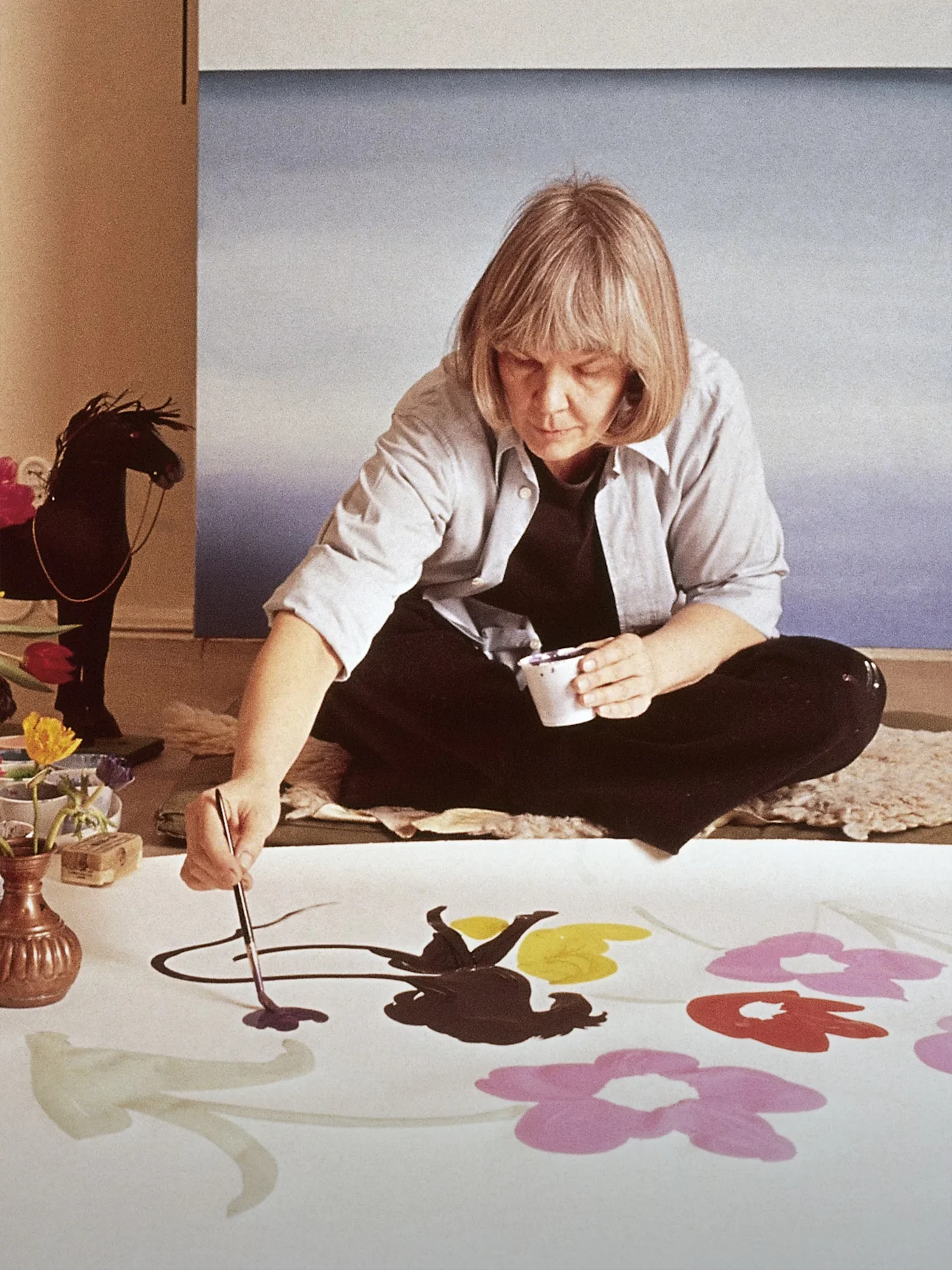 Artystka Marimekko Maija Isola siedzi na podłodze, malując duże, kolorowe kwiaty na białym materiale.