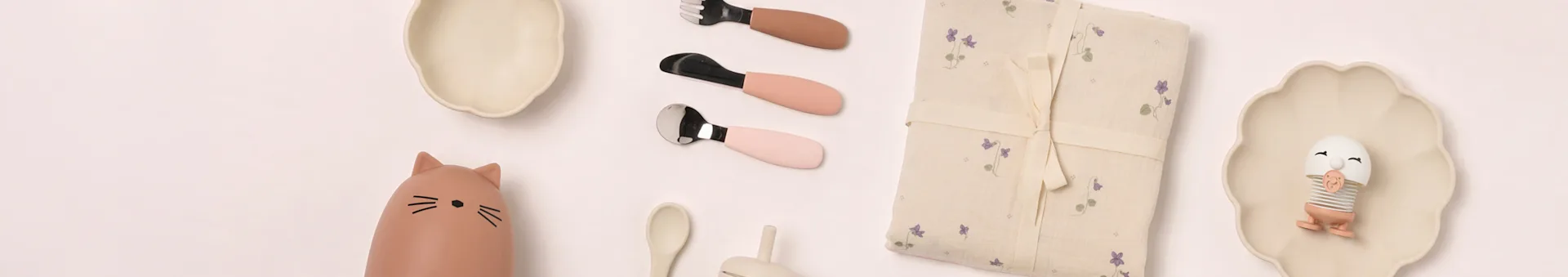 Artykuły dziecięce flat lay: beżowe talerze, butelka kot, różowe sztućce, koc w kwiaty, zabawka jajko.